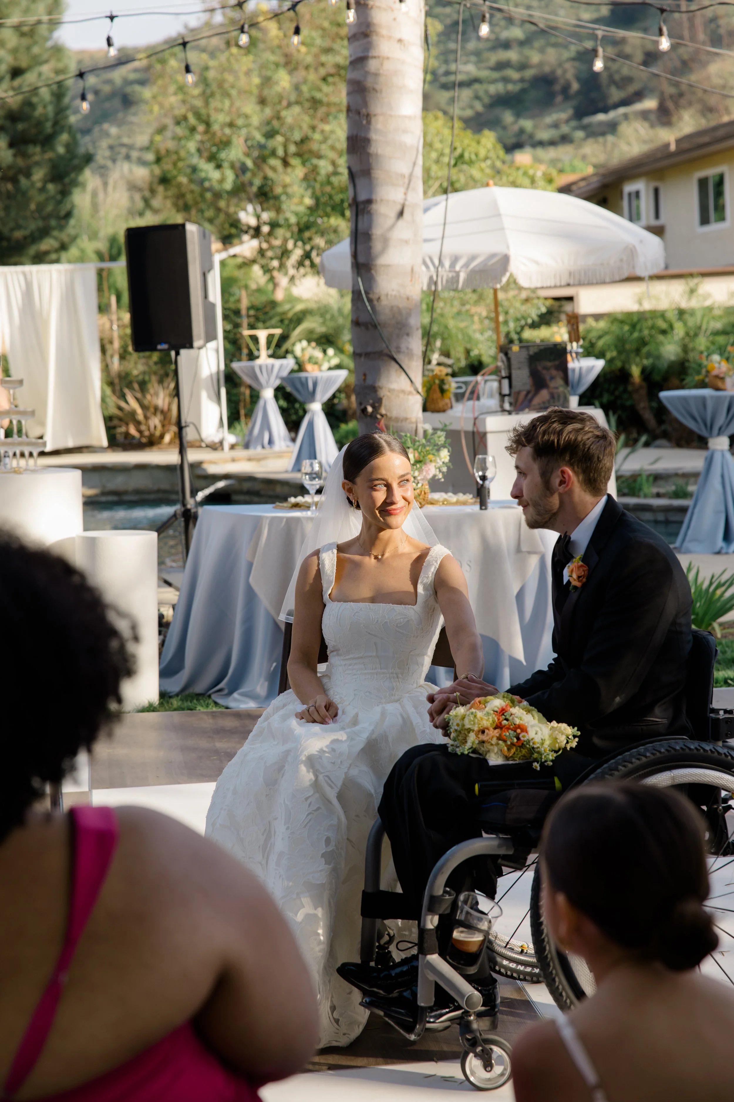 A bride and groom sitting together during their outdoor wedding ceremony. The bride is wearing a white wedding dress and veil, holding a small bouquet, while the groom is in a black suit with a boutonnière. Guests are seated in front, with a diverse 