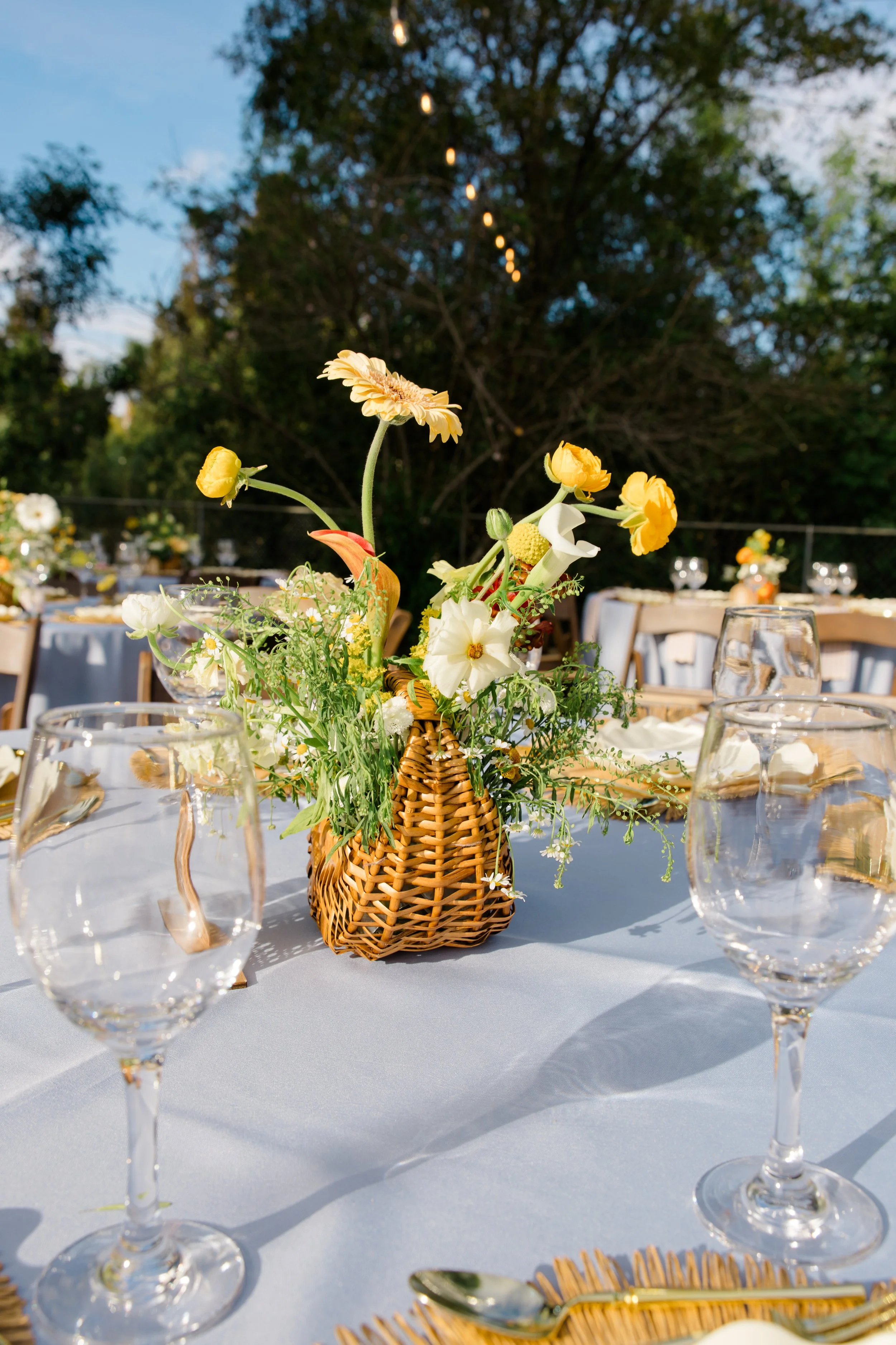 Outdoor dining table with a central floral arrangement in a wicker basket, surrounded by empty wine glasses, on a white tablecloth, with trees and string lights visible in the background during the day.