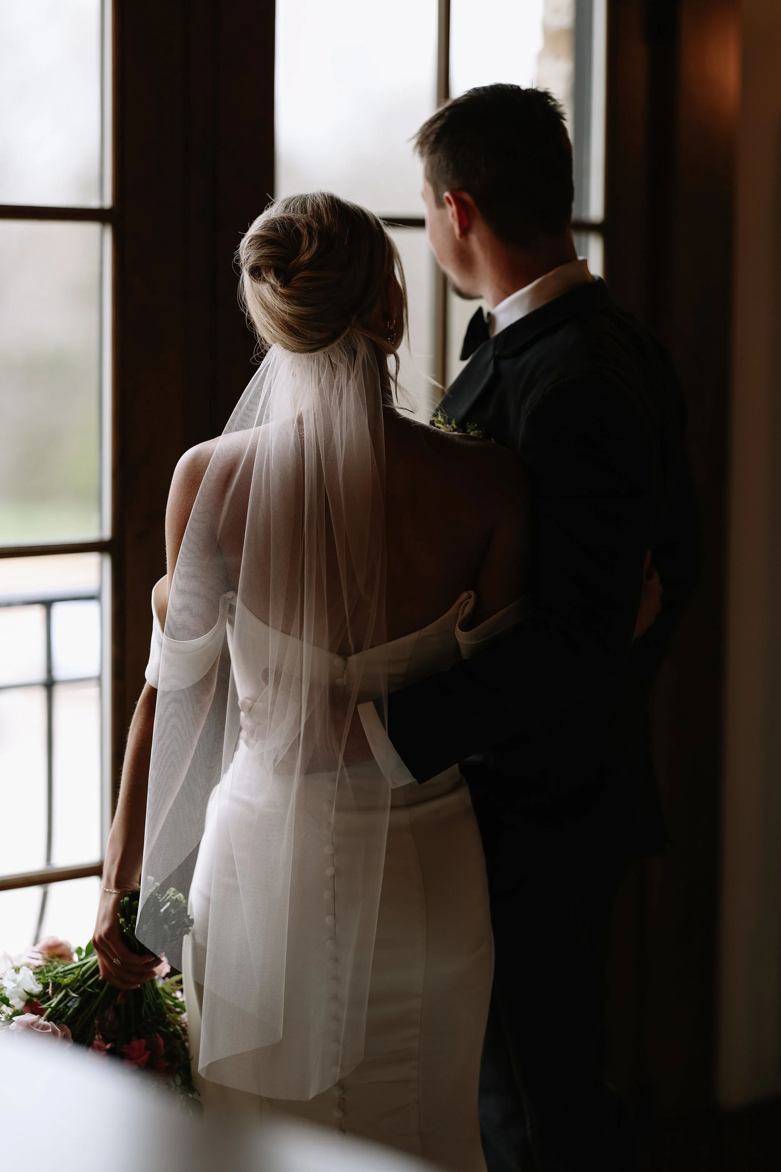 A bride and groom standing close together indoors near a window, with the bride holding a bouquet of flowers.