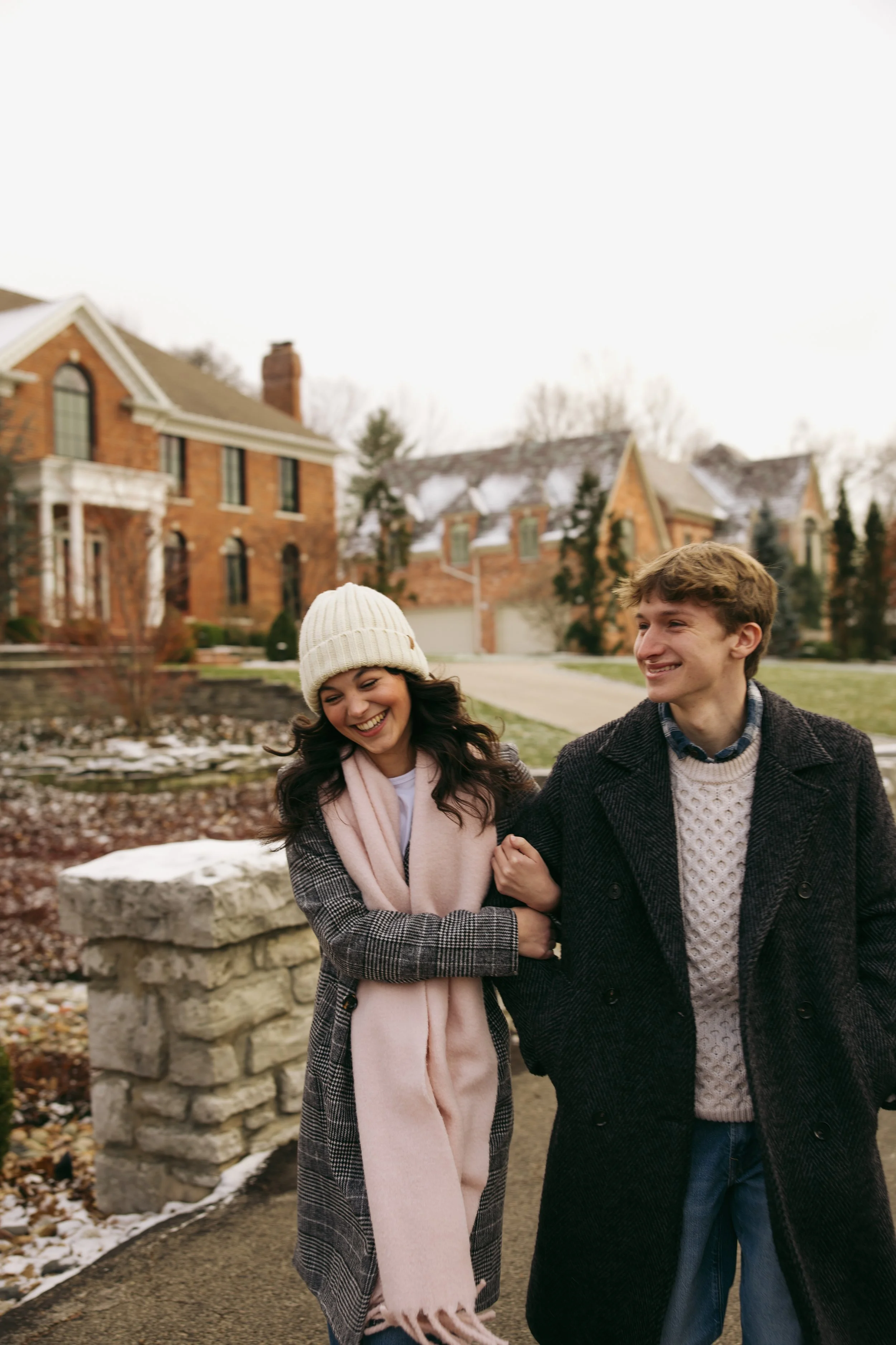 A young woman and man are smiling and holding hands outside in a neighborhood. The woman is wearing a beige hat, a pink scarf, and a patterned coat. The man is wearing a dark coat and a light-colored sweater. There are houses and trees in the backgro