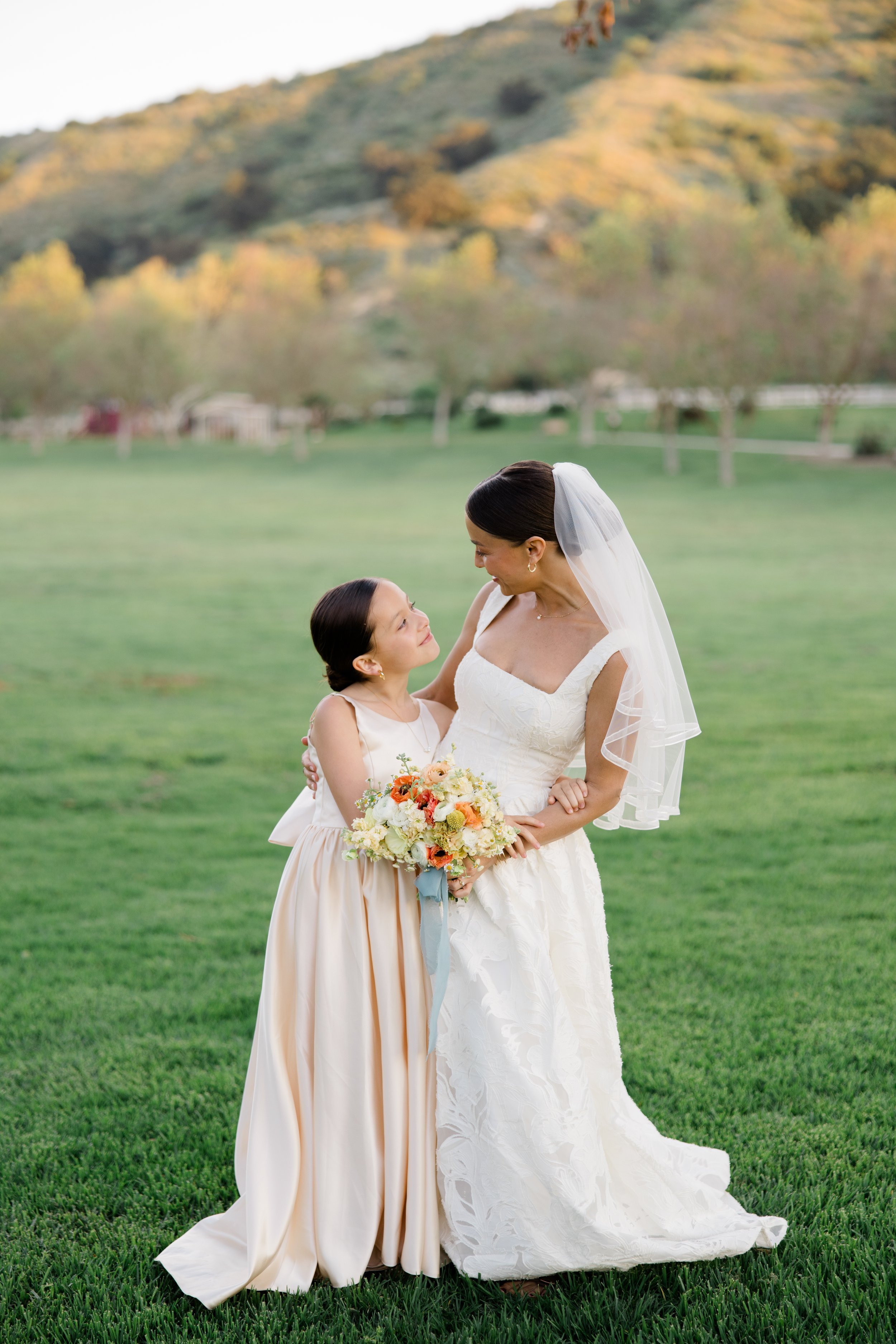 A bride in a white wedding dress holding a bouquet, standing on grass and looking at a young girl in a beige dress, with landscape of trees and hills in the background.