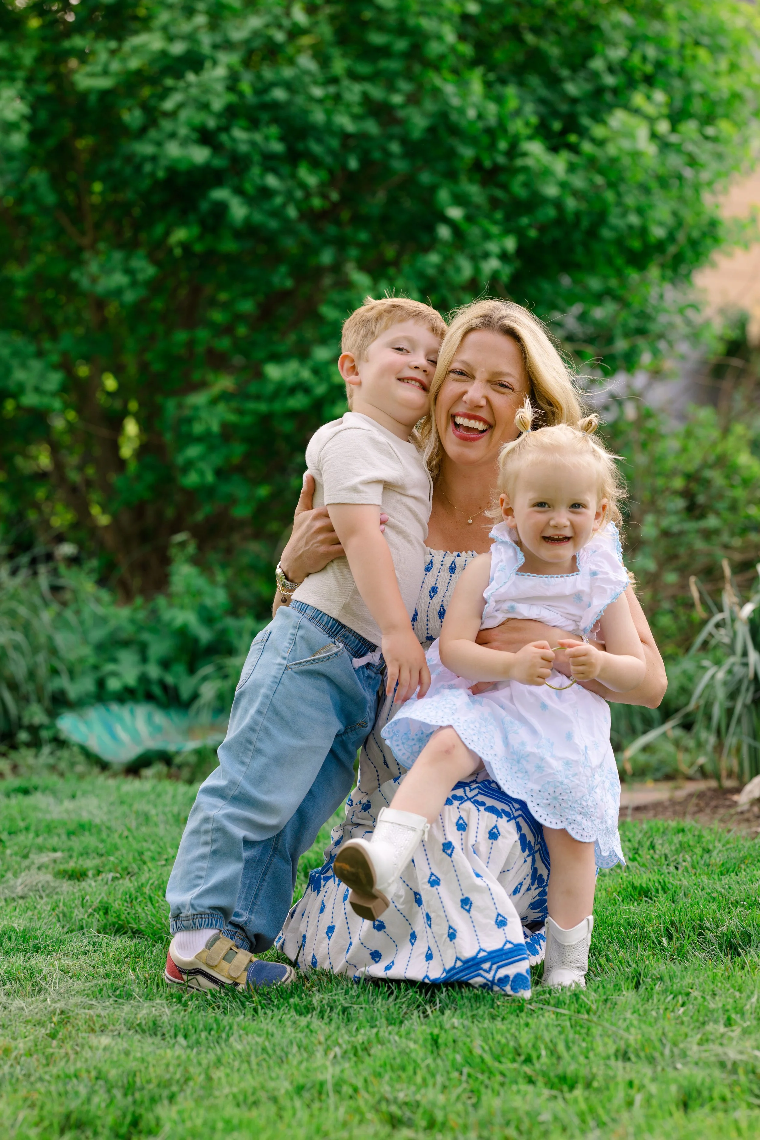 A woman smiling and holding two young children outdoors in a garden with green trees and grass.