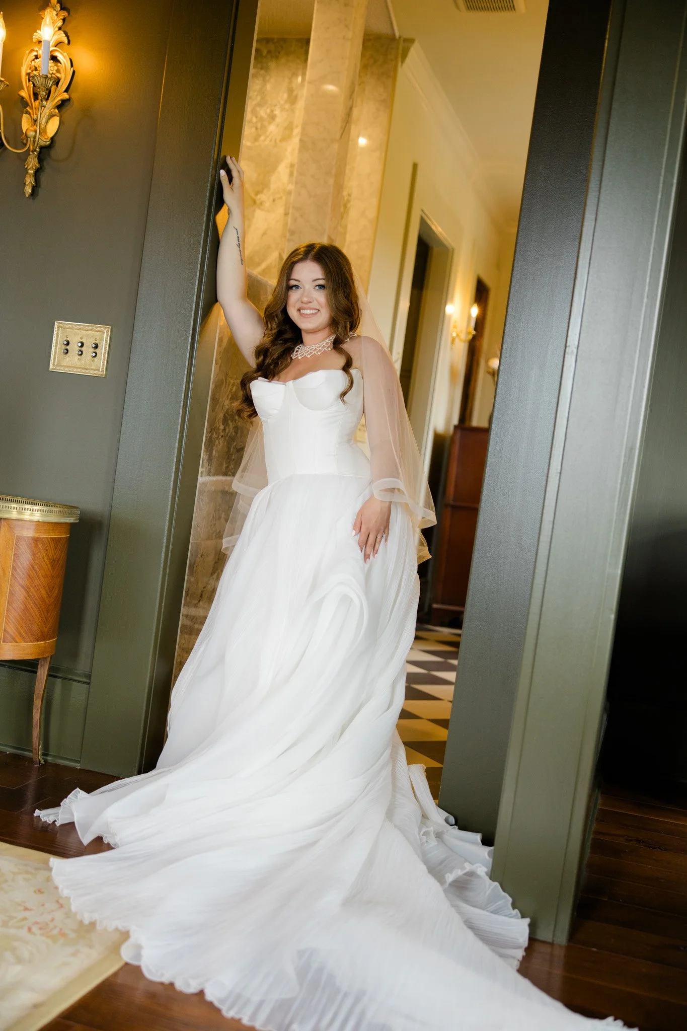 A smiling woman in a white wedding dress with long flowing skirt, standing in a hallway with elegant lighting and marble walls.
