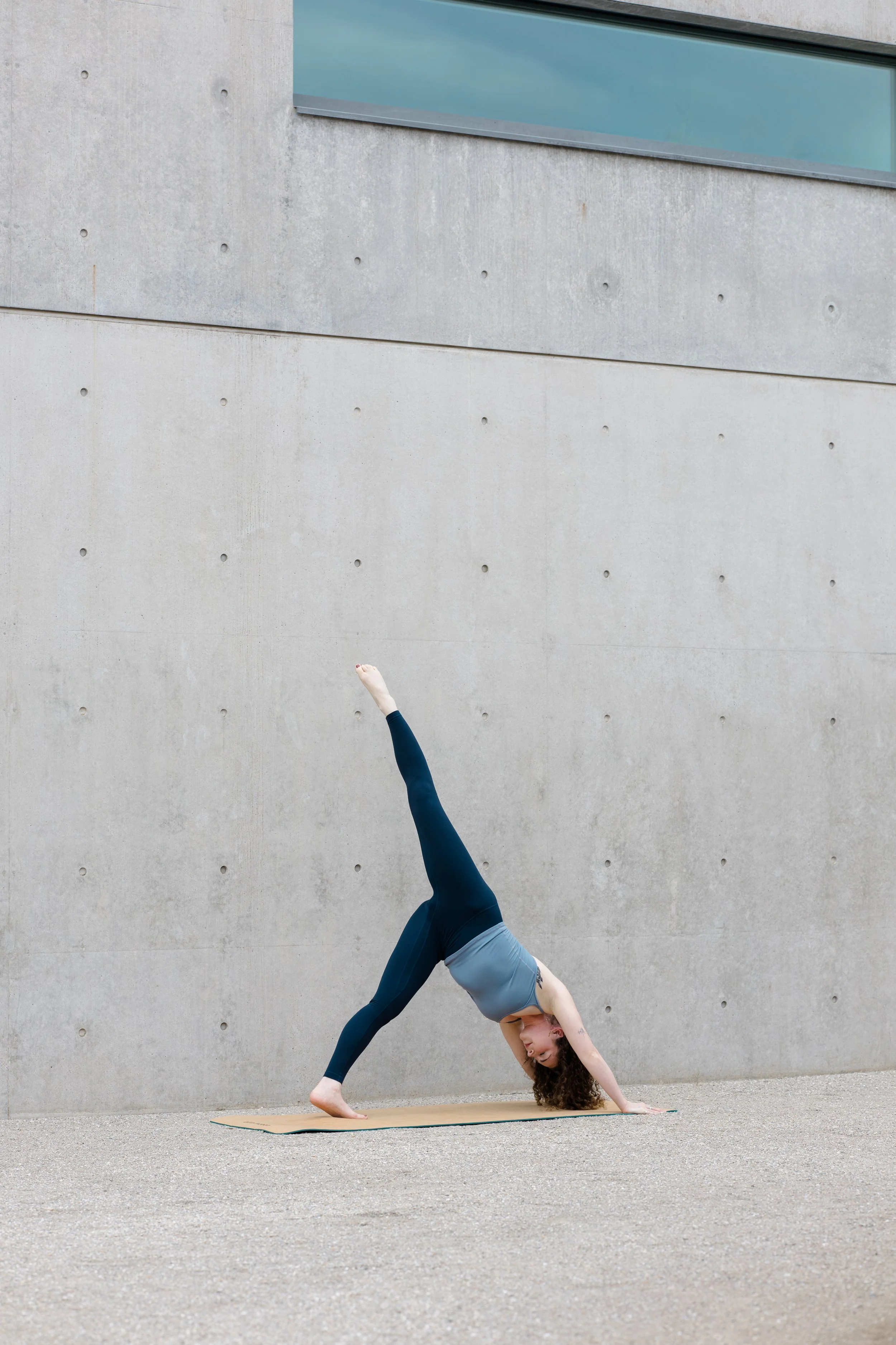 A woman practicing yoga outdoors on a mat, performing a handstand with her legs extended against a concrete wall.