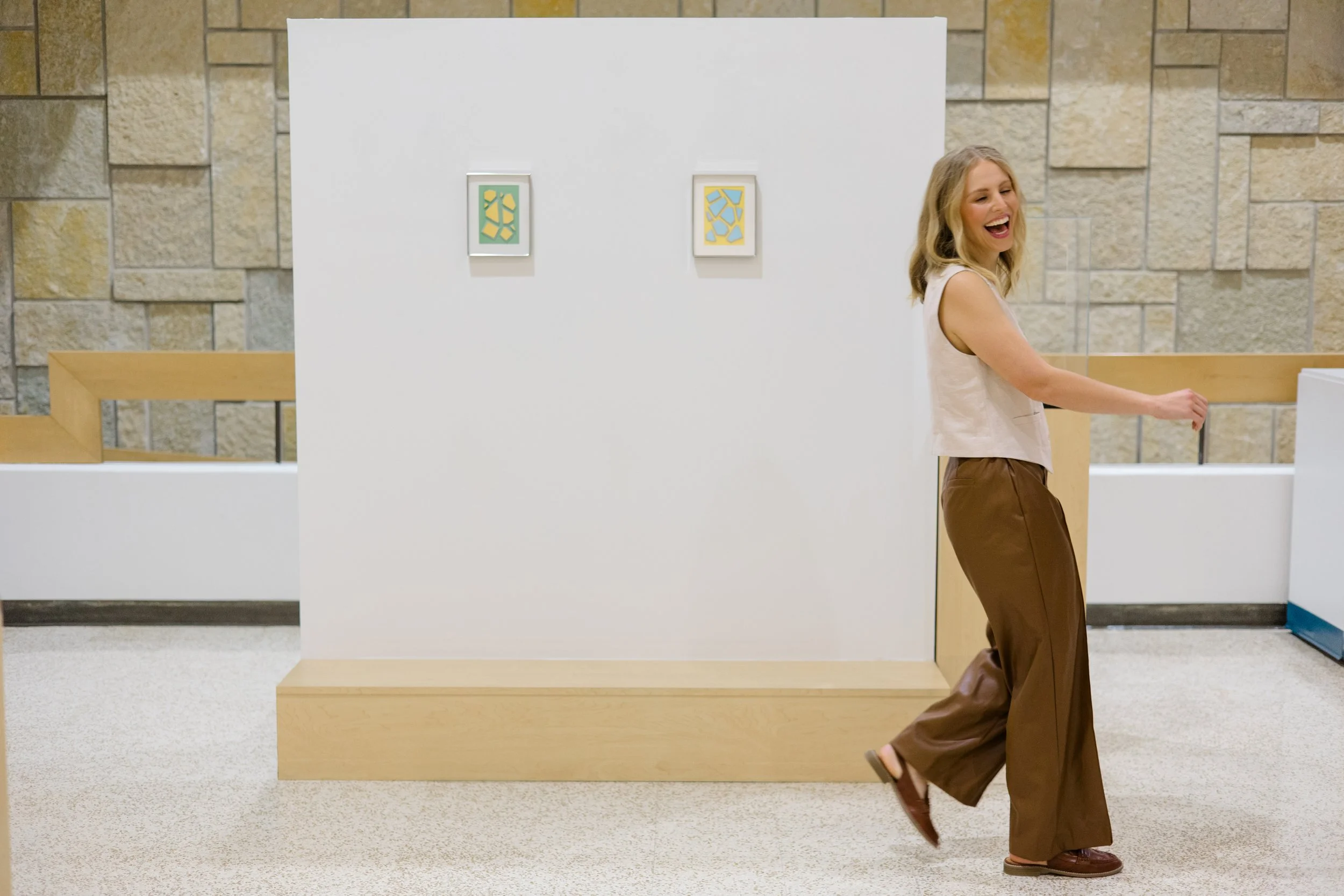 A woman walking past a white partition at an art gallery, with abstract artwork hanging on the partition, and a stone wall in the background.