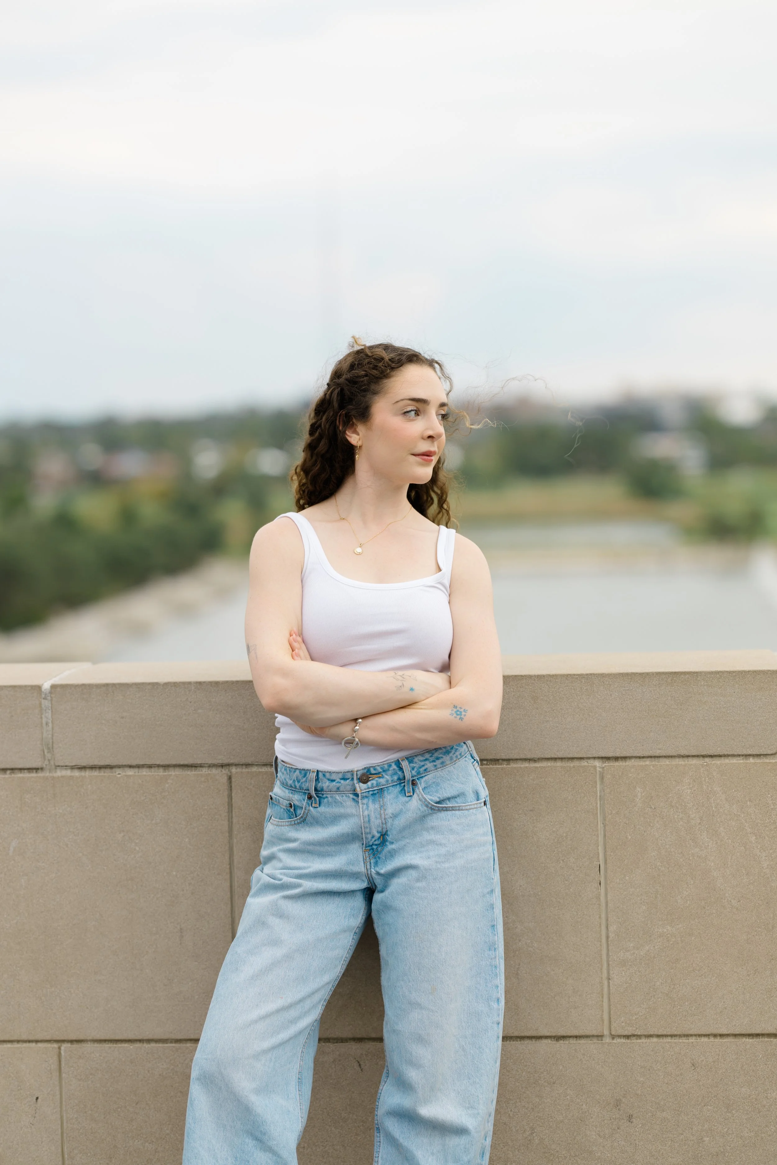 A young woman with curly brown hair standing outdoors with arms crossed, wearing a white tank top, blue jeans, and jewelry, against a background of a river and distant trees under a cloudy sky.