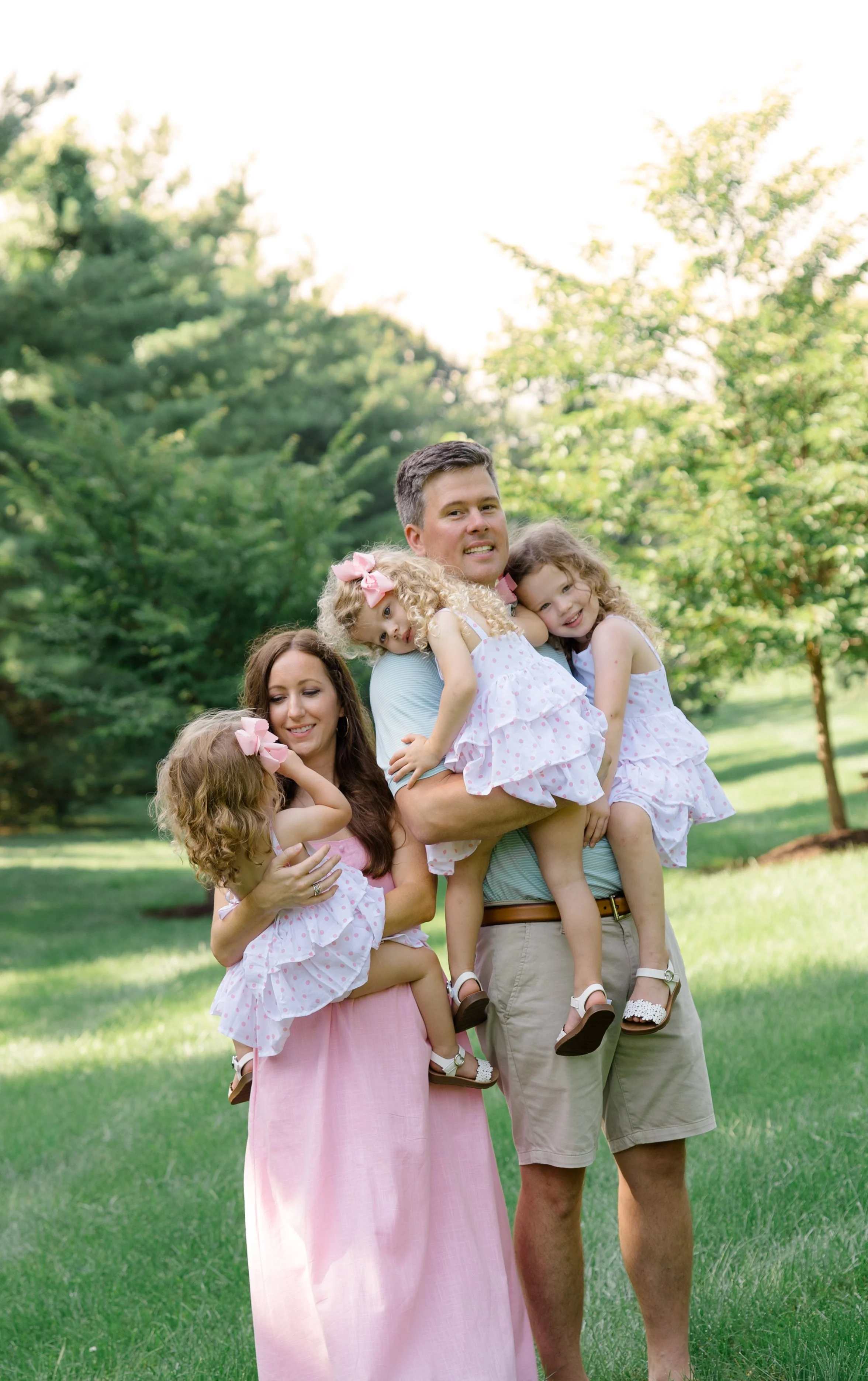 A family group outdoors on a grassy area with trees in the background, includes a man and woman holding three young girls, all dressed in light-colored summer clothes and smiling.