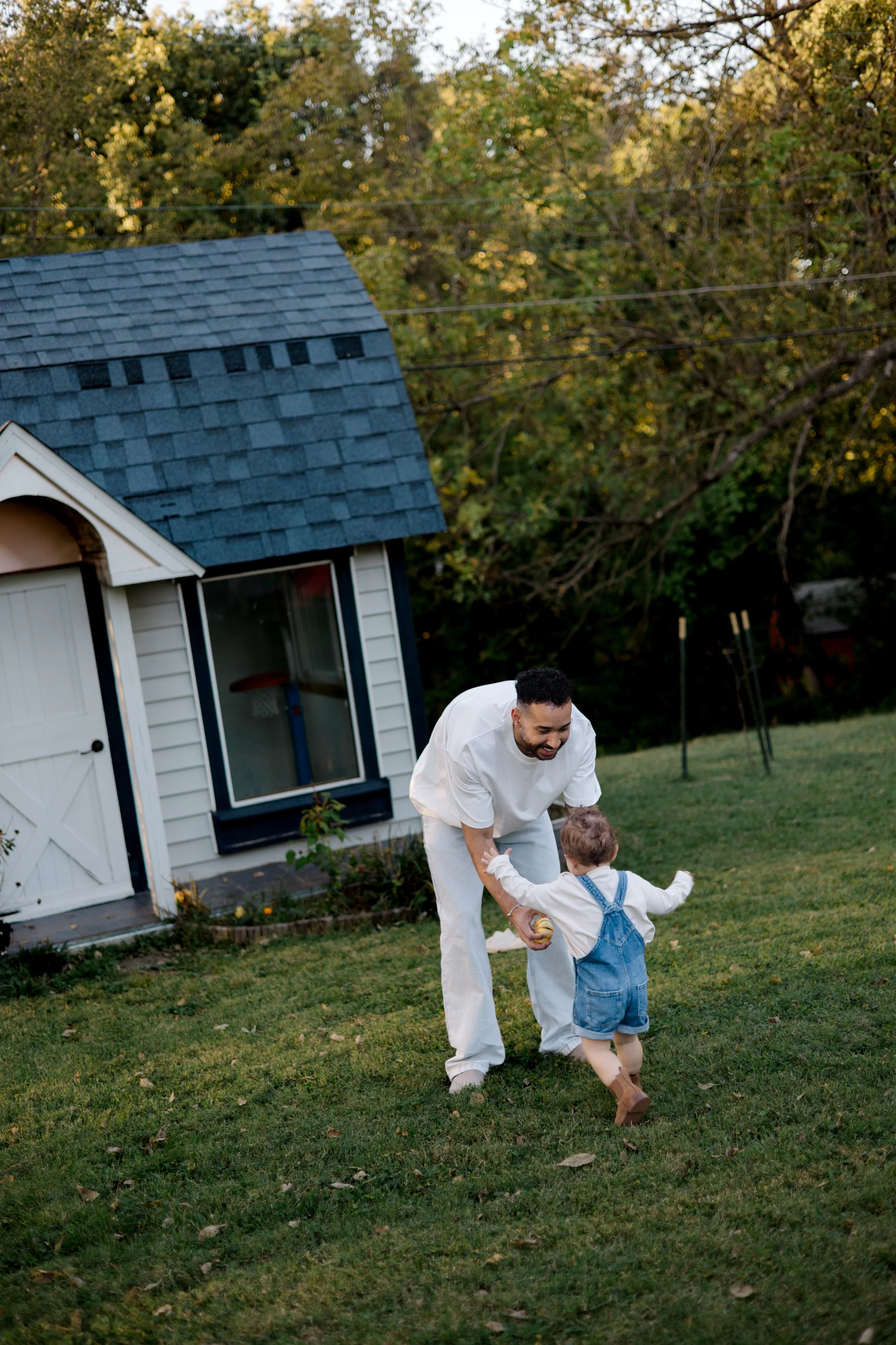 An adult man playing with a young child in a backyard yard, with a small white shed with black trim and trees in the background.