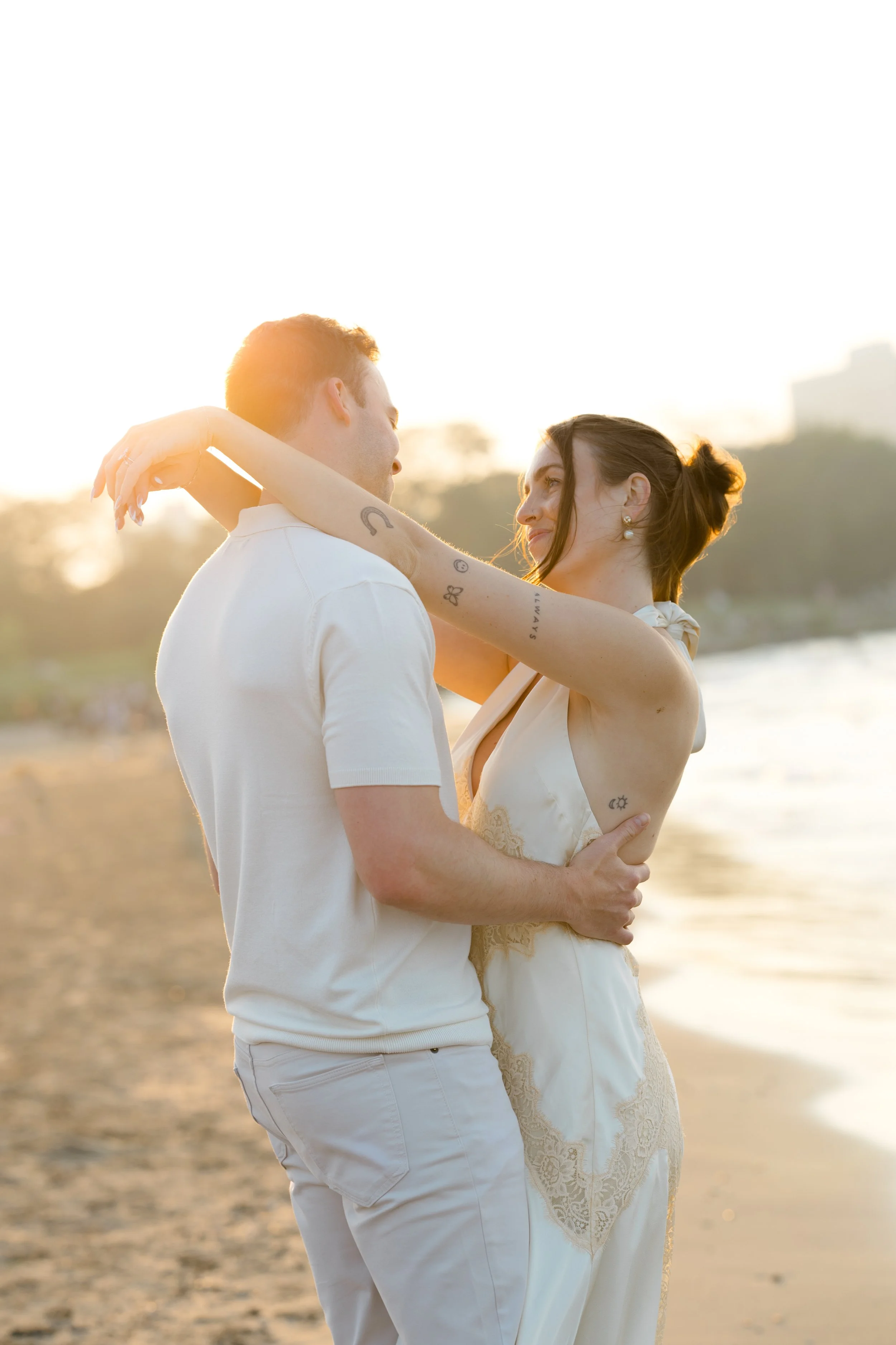 A couple dancing closely on a beach during sunset, with the man wearing a white shirt and the woman in a white dress, both showing affectionate expressions.