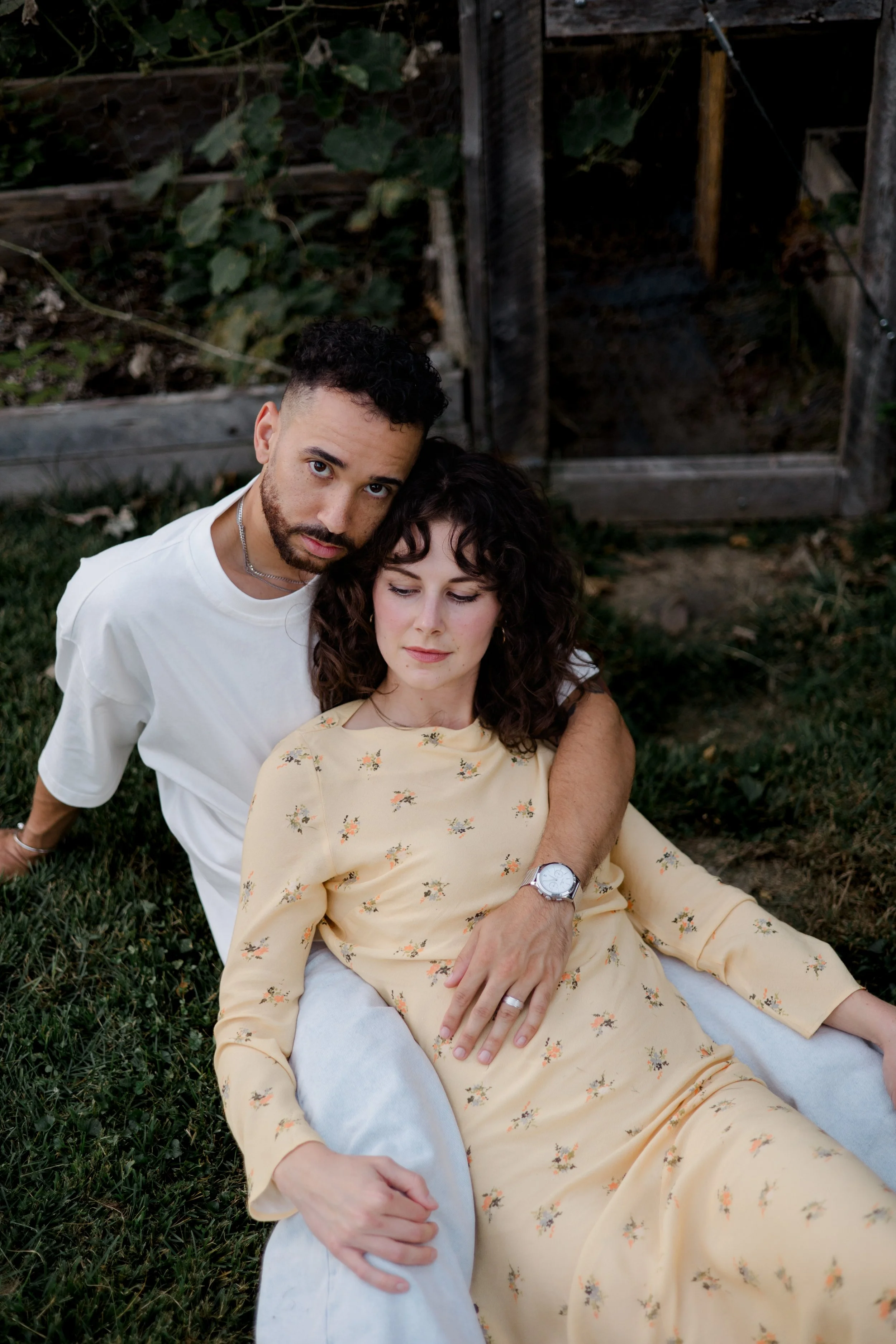 A man and woman sitting on grass in front of a wooden fence with greenery. The man has short curly hair, a beard, and is wearing a white shirt. The woman has curly hair, is wearing a yellow floral dress, and has a wristwatch and rings. The man looks 