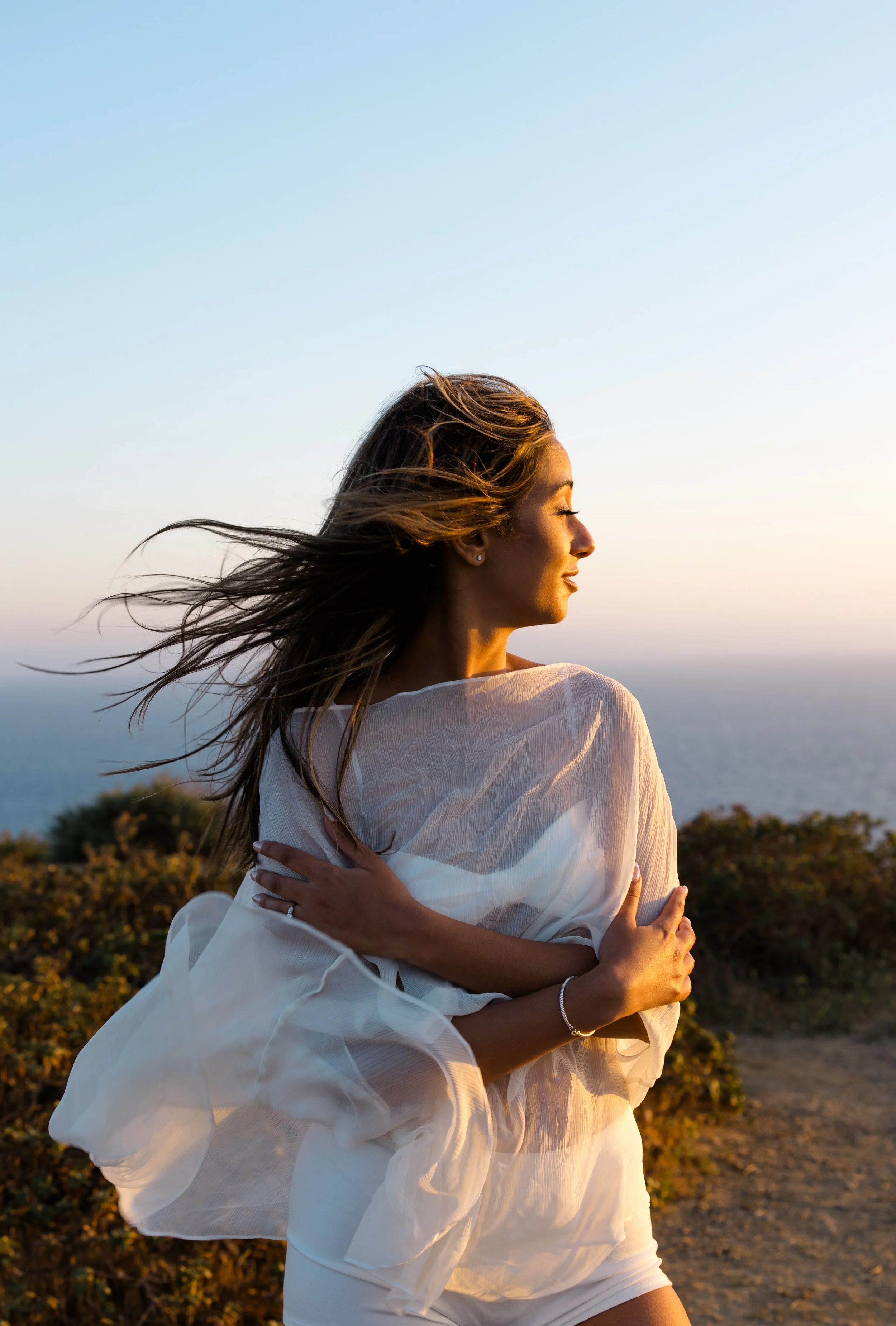 A woman with long, wind-blown hair standing outdoors at sunset, wearing a white dress with her arms crossed over her chest, looking peacefully to the side.