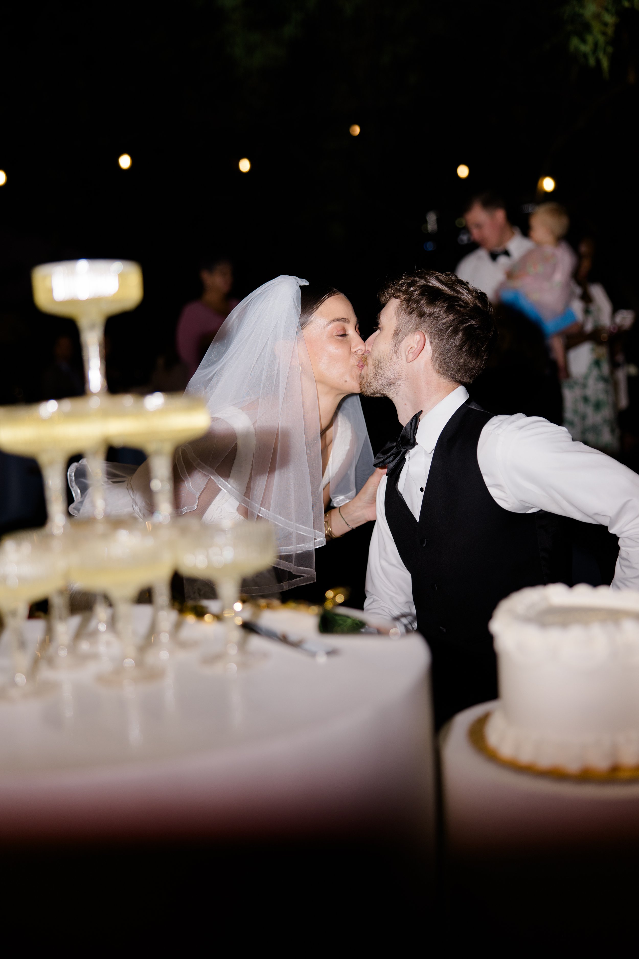 A bride and groom kissing at their wedding reception, with blurry guests and wedding cake in the background.