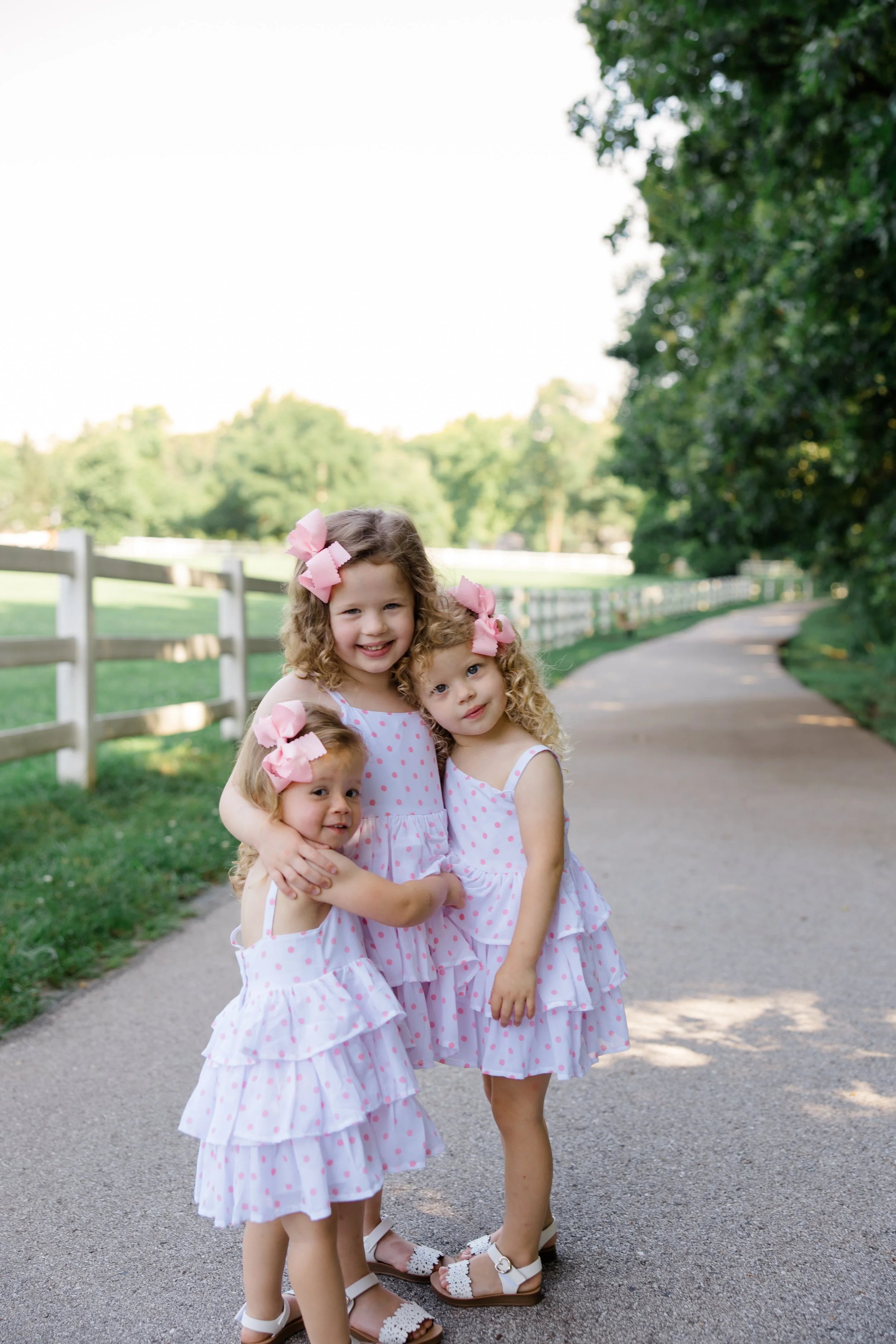 Four young girls with curly hair and pink bows in their hair, wearing matching pink polka-dot dresses, standing on a paved path in a park, hugging each other and smiling.