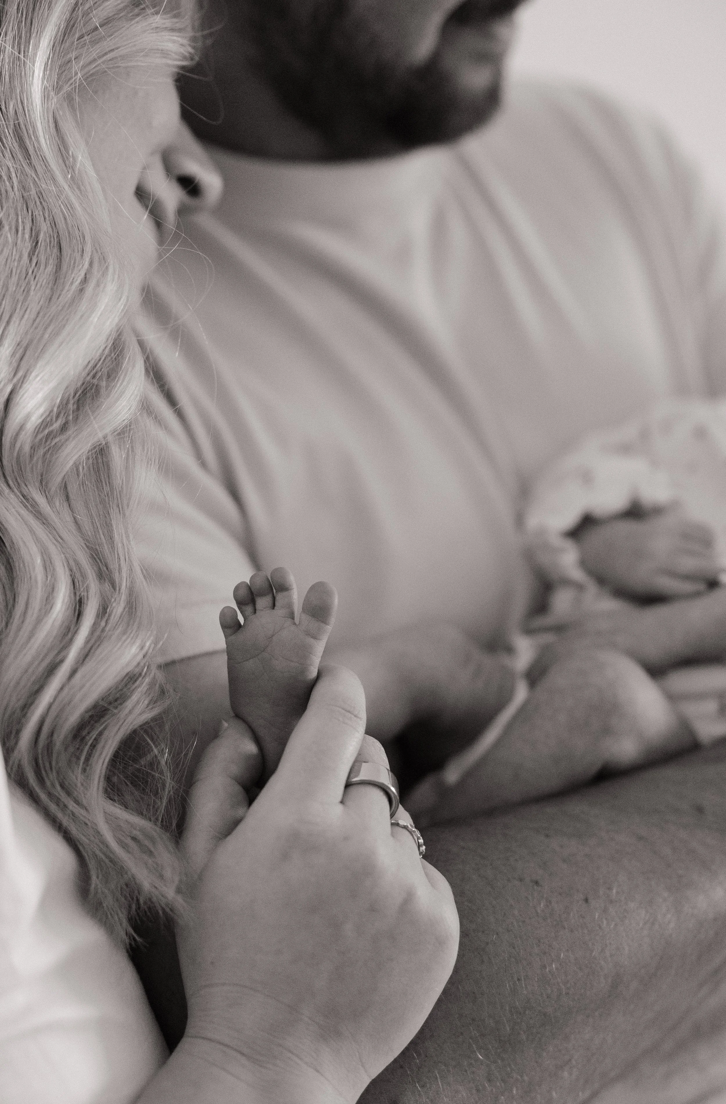 Close-up of a woman with long, wavy hair holding a baby's hand. The woman is wearing rings and a bracelet, and the scene appears tender and intimate.