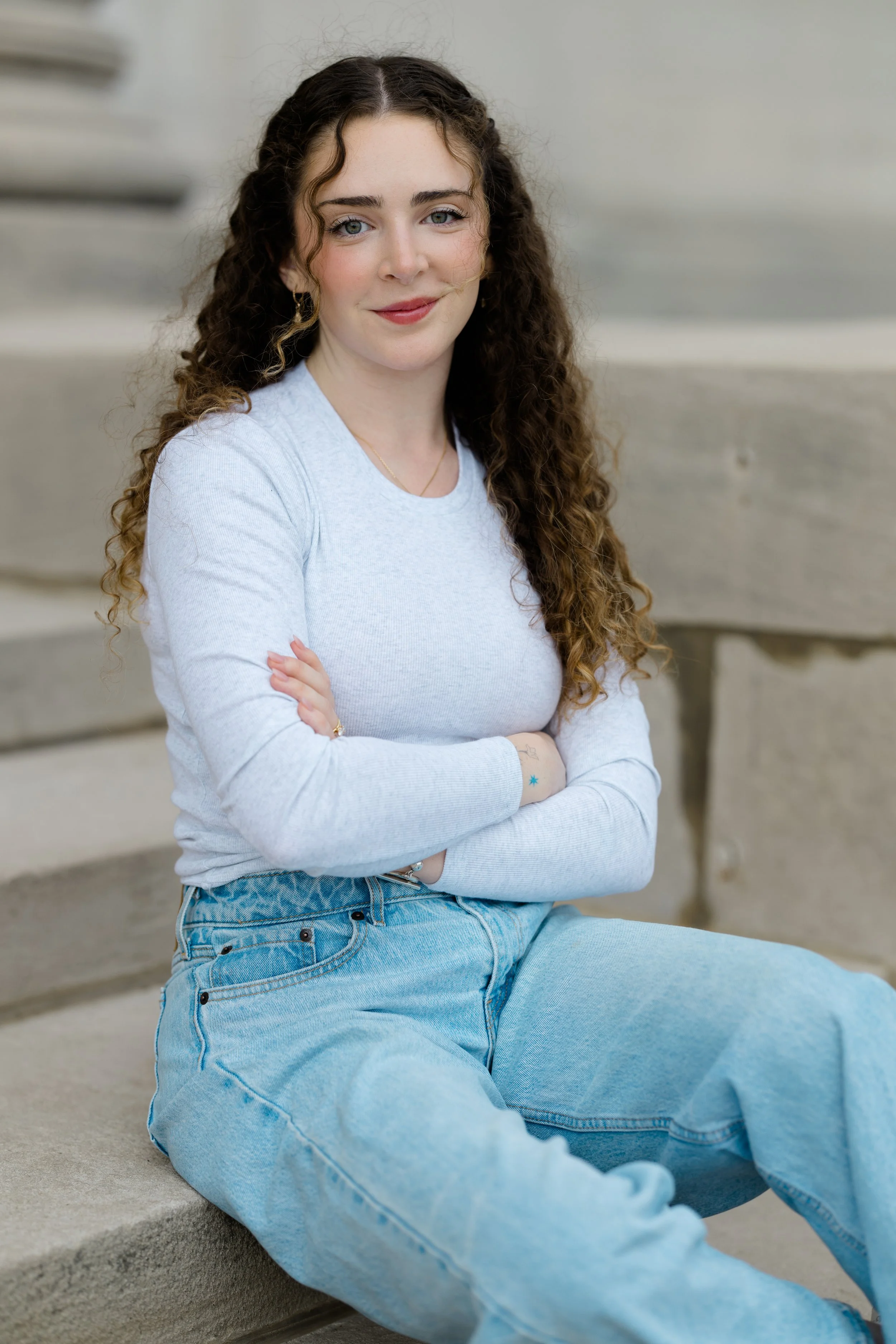 A young woman with curly brown hair, blue eyes, and light skin, sitting on concrete steps, wearing a light gray long-sleeve shirt and blue jeans, smiling softly.
