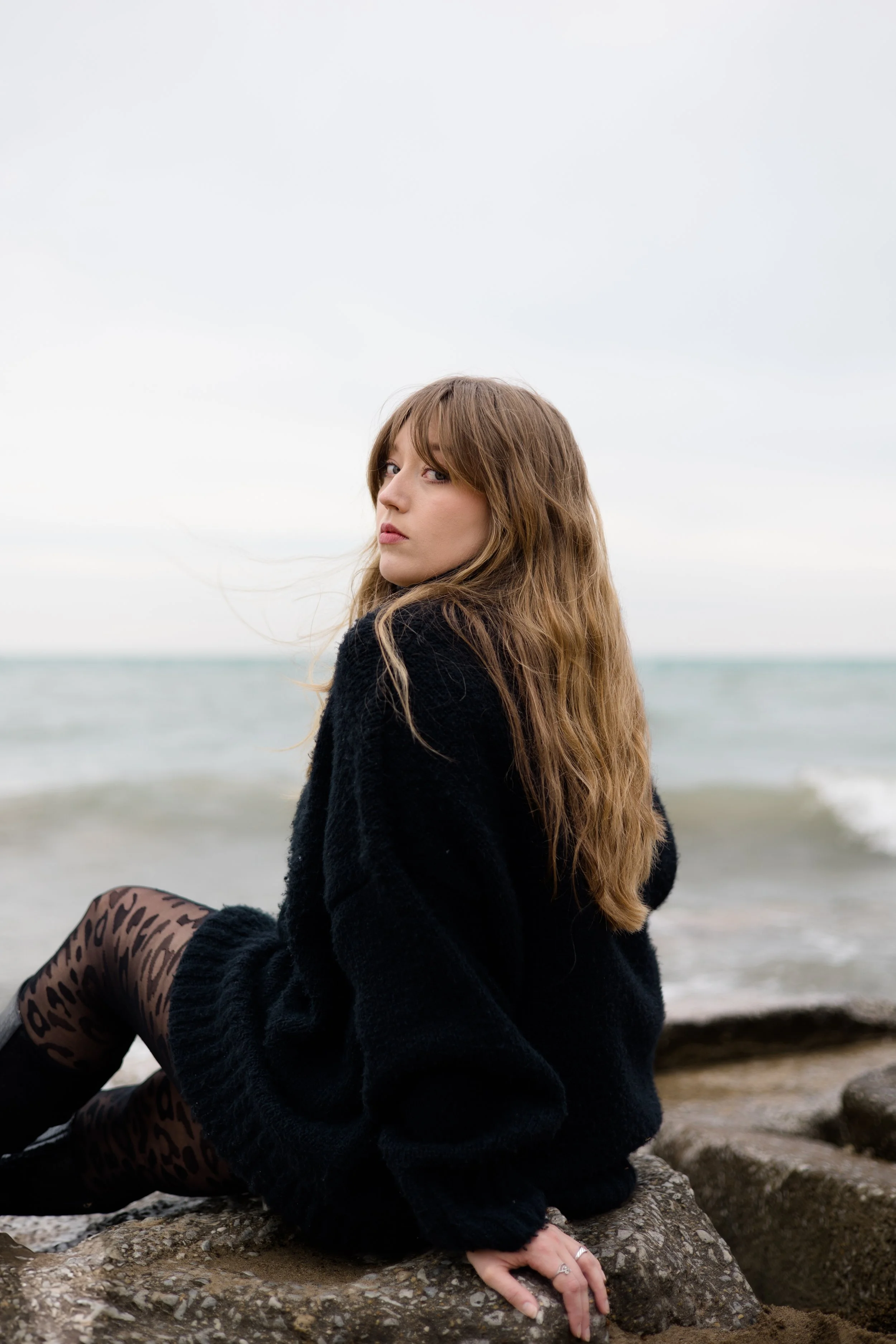A woman with long brown hair sitting on rocks by the beach, wearing a black fuzzy coat and leopard print leggings, with the ocean and sky in the background.