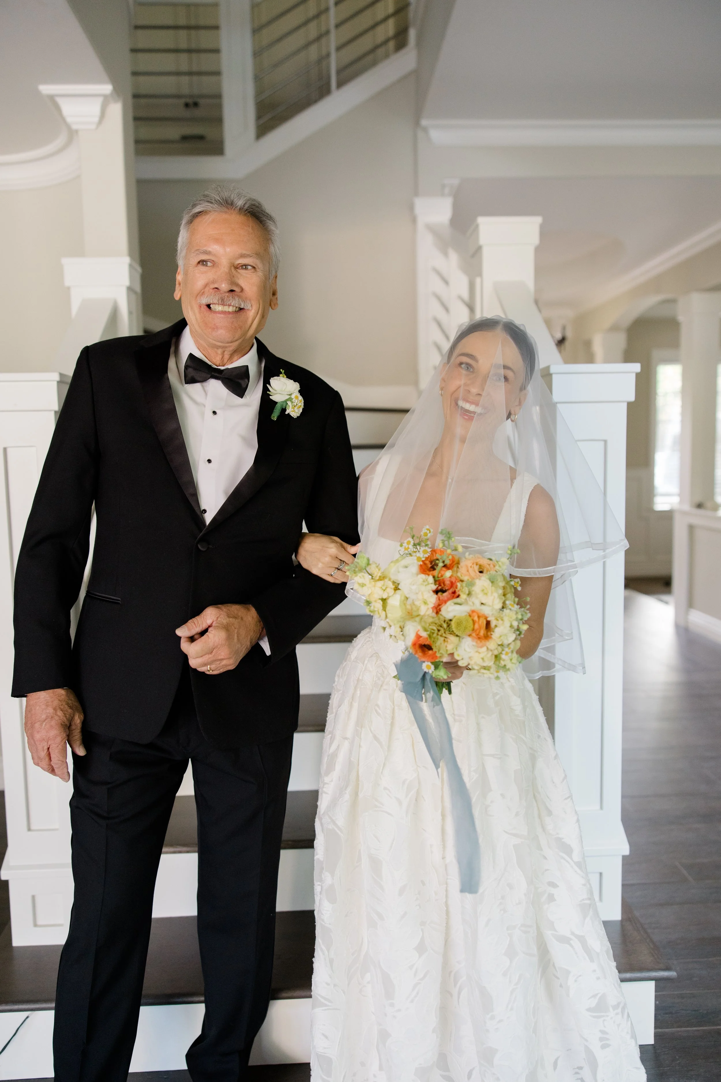 An elderly man in a tuxedo and a white boutonniere standing next to a smiling bride with a veil, holding a bouquet of flowers inside a house.