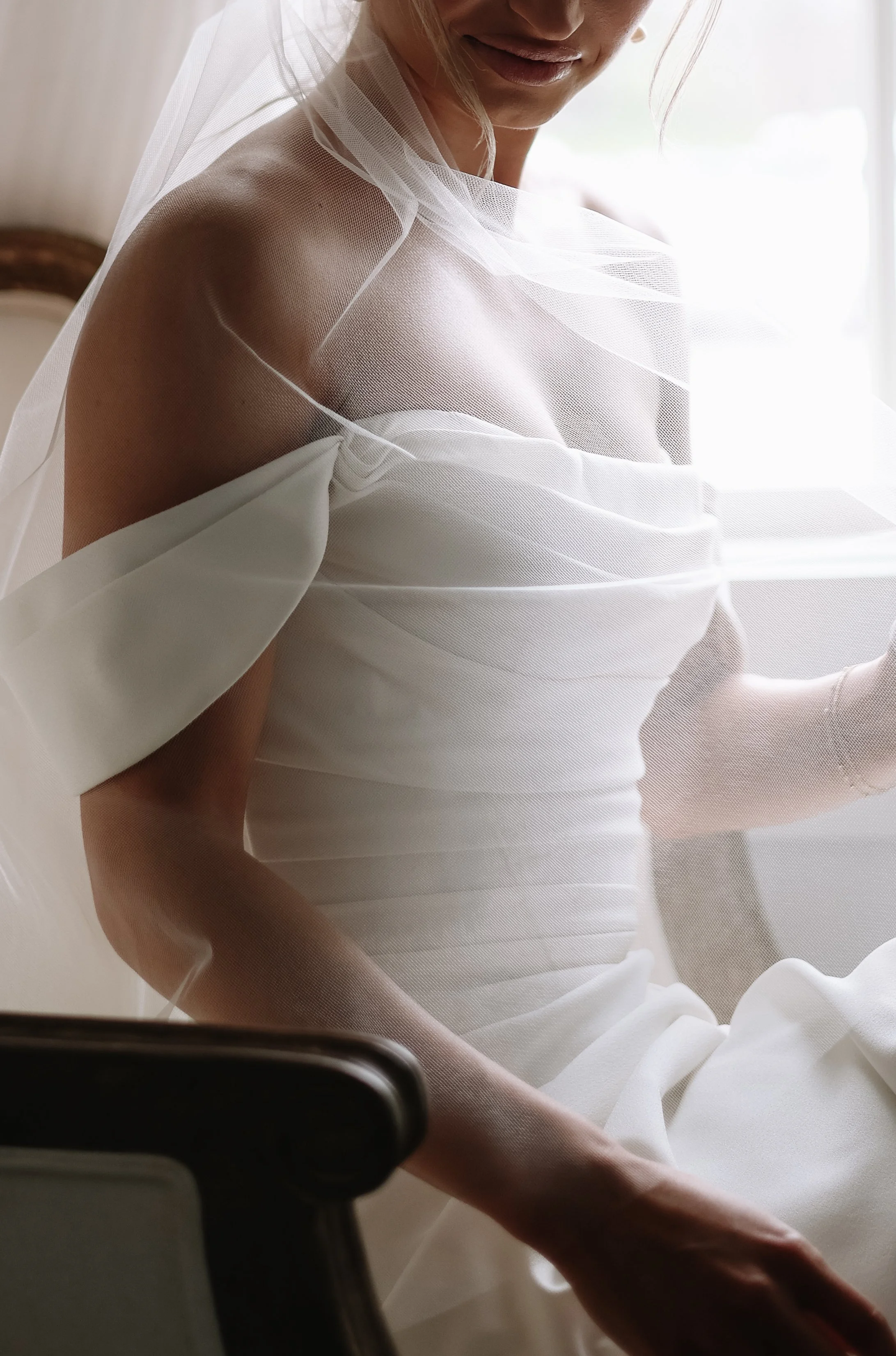 A woman dressed in white, sitting near a window with soft natural light, partially covered by sheer white fabric, with her face slightly turned and a gentle expression.