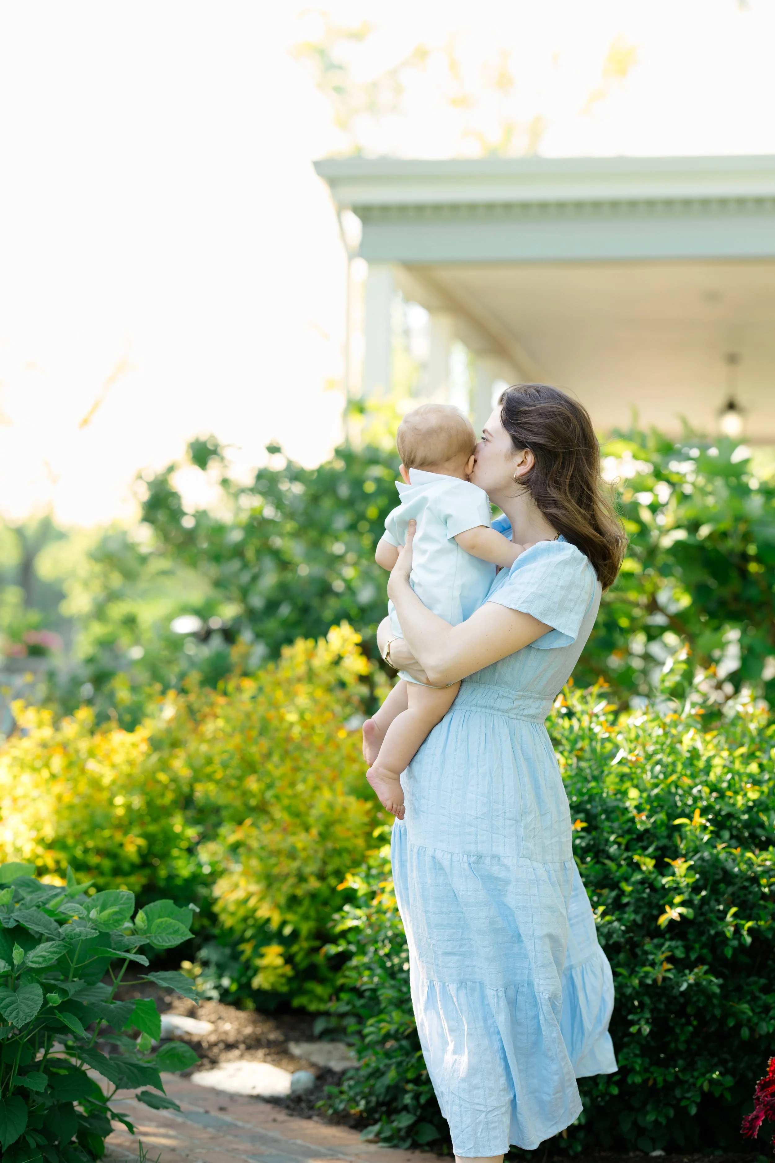 A woman holding and kissing a young child in a garden with lush green plants and a house in the background.