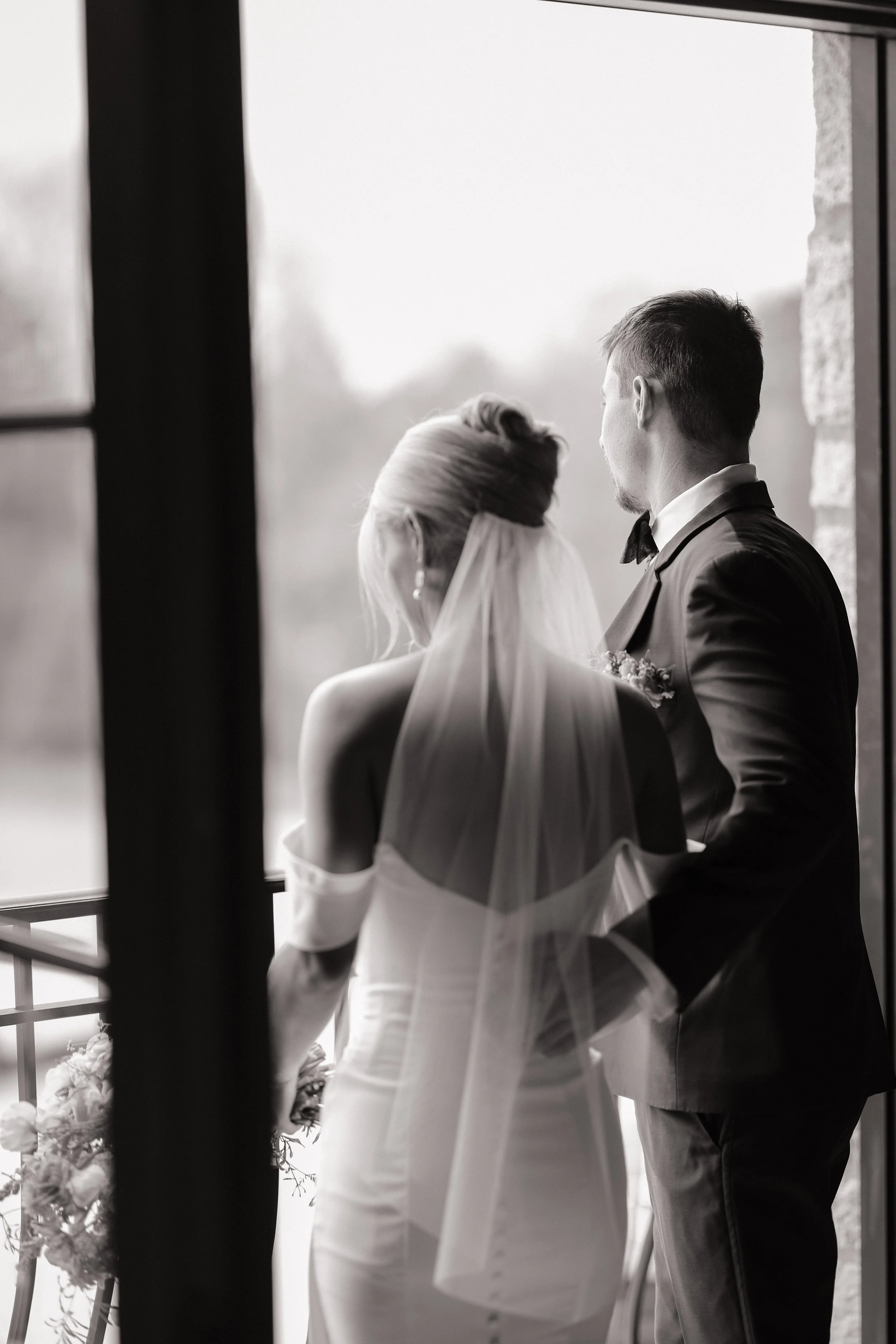 A black and white photo of a groom and bride standing near a window, seen from behind, during a wedding ceremony.