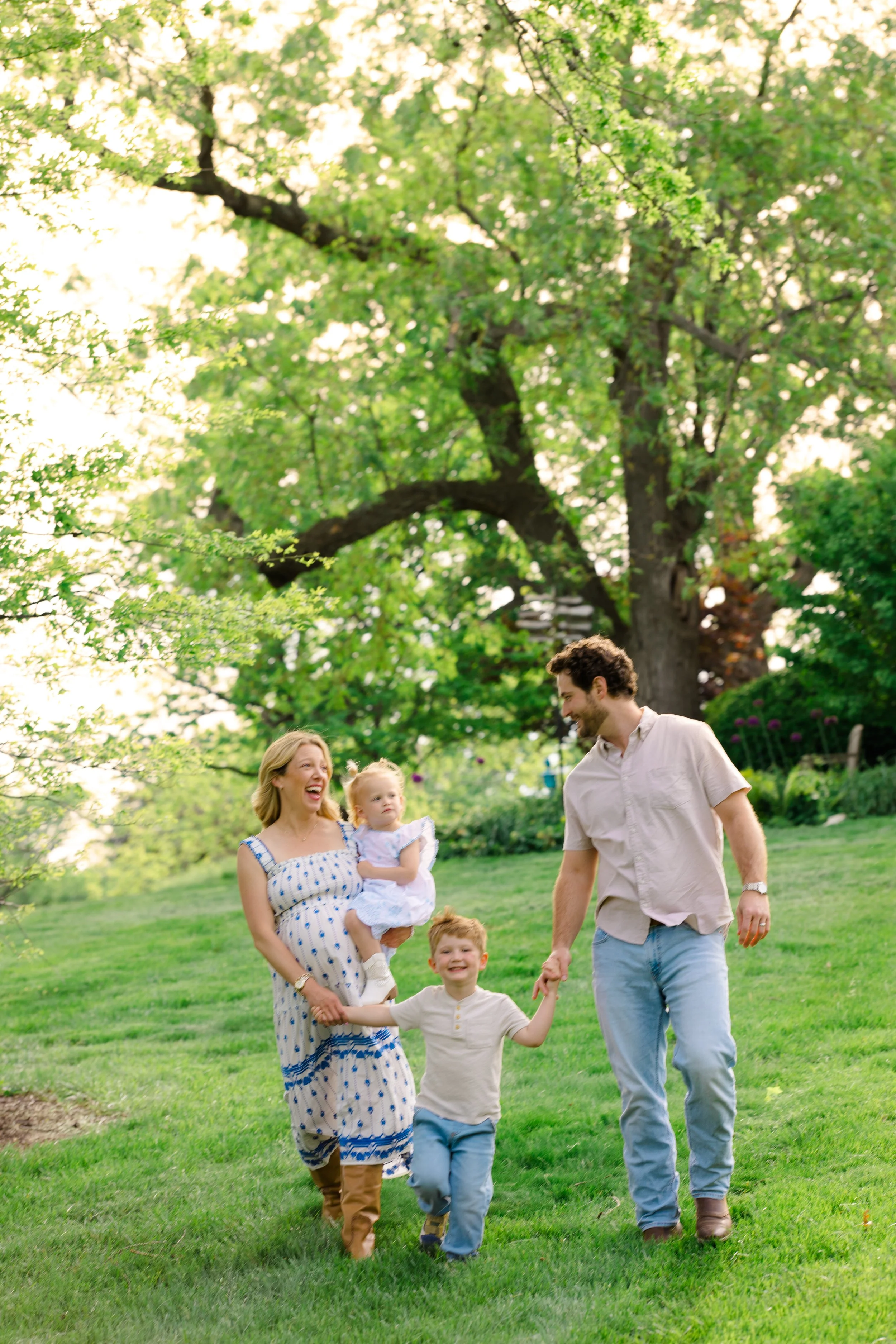 A family of four enjoying a walk together in a park with green grass and trees, smiling and holding hands.