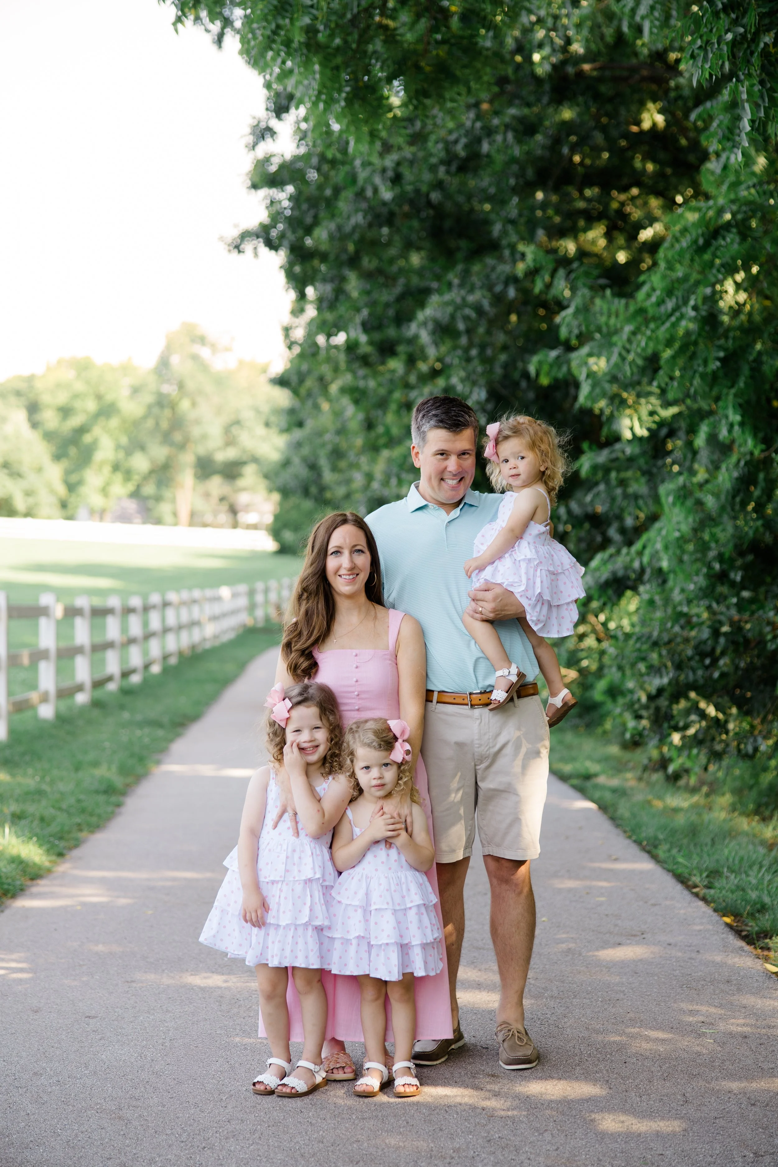A family of five, including a father, mother, and three young daughters, standing on a sidewalk with lush green trees and a grassy park in the background, all dressed in light pink and white outfits.
