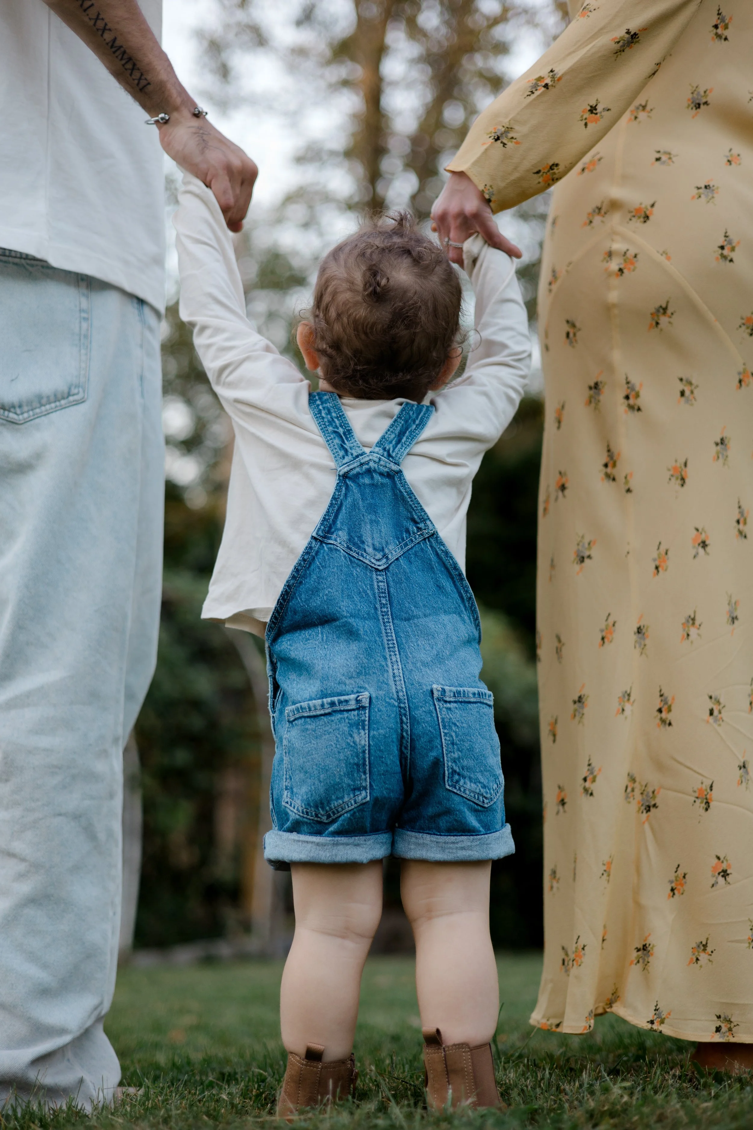 A young child holding hands with two adults outdoors, with a background of trees and grass. The child is wearing denim overalls and brown boots.