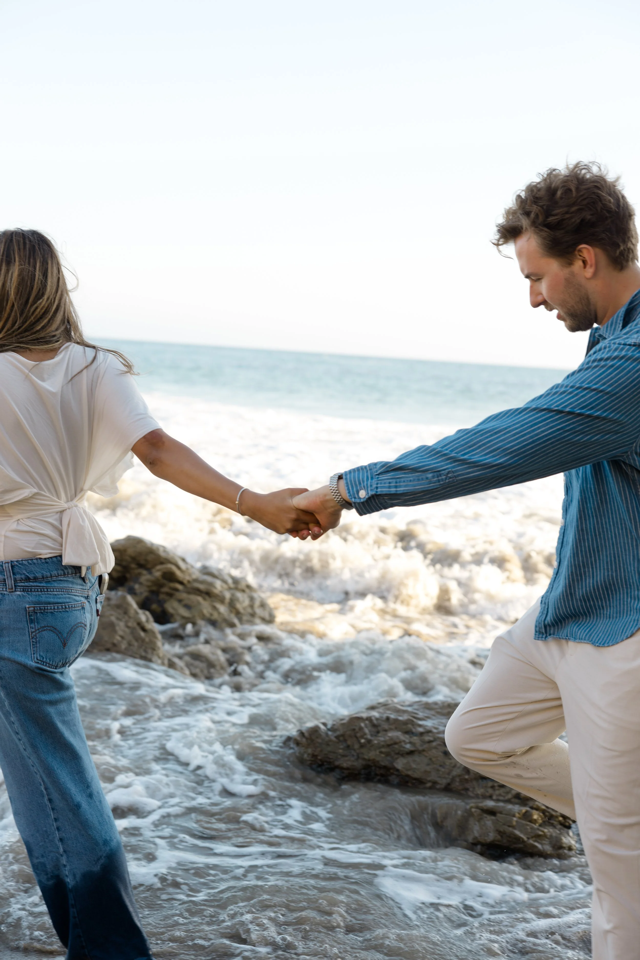 A couple holding hands on the beach with ocean waves, rocks, and sky in the background.