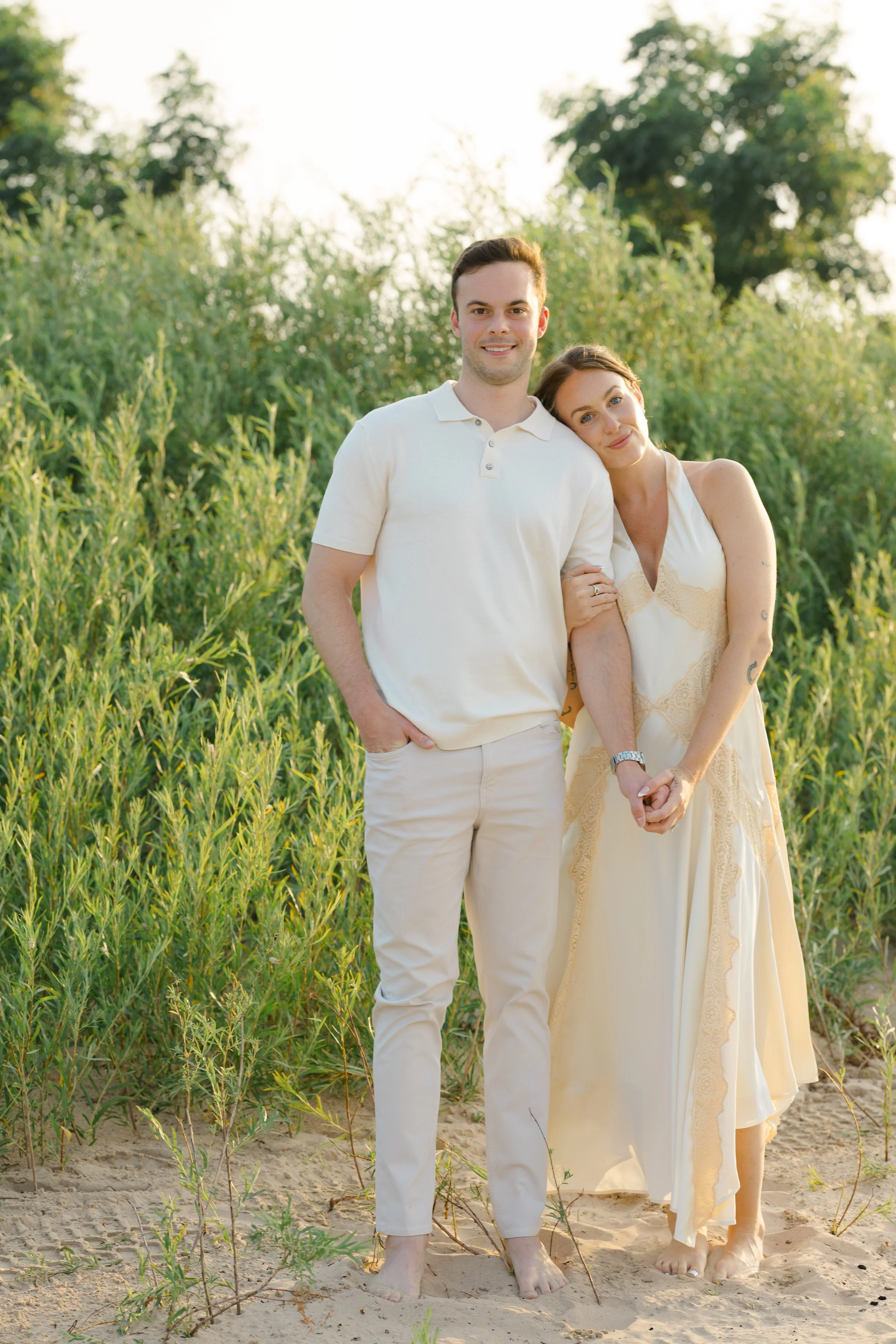 A couple standing close together on a sandy beach with lush greenery and trees behind them, during sunset.