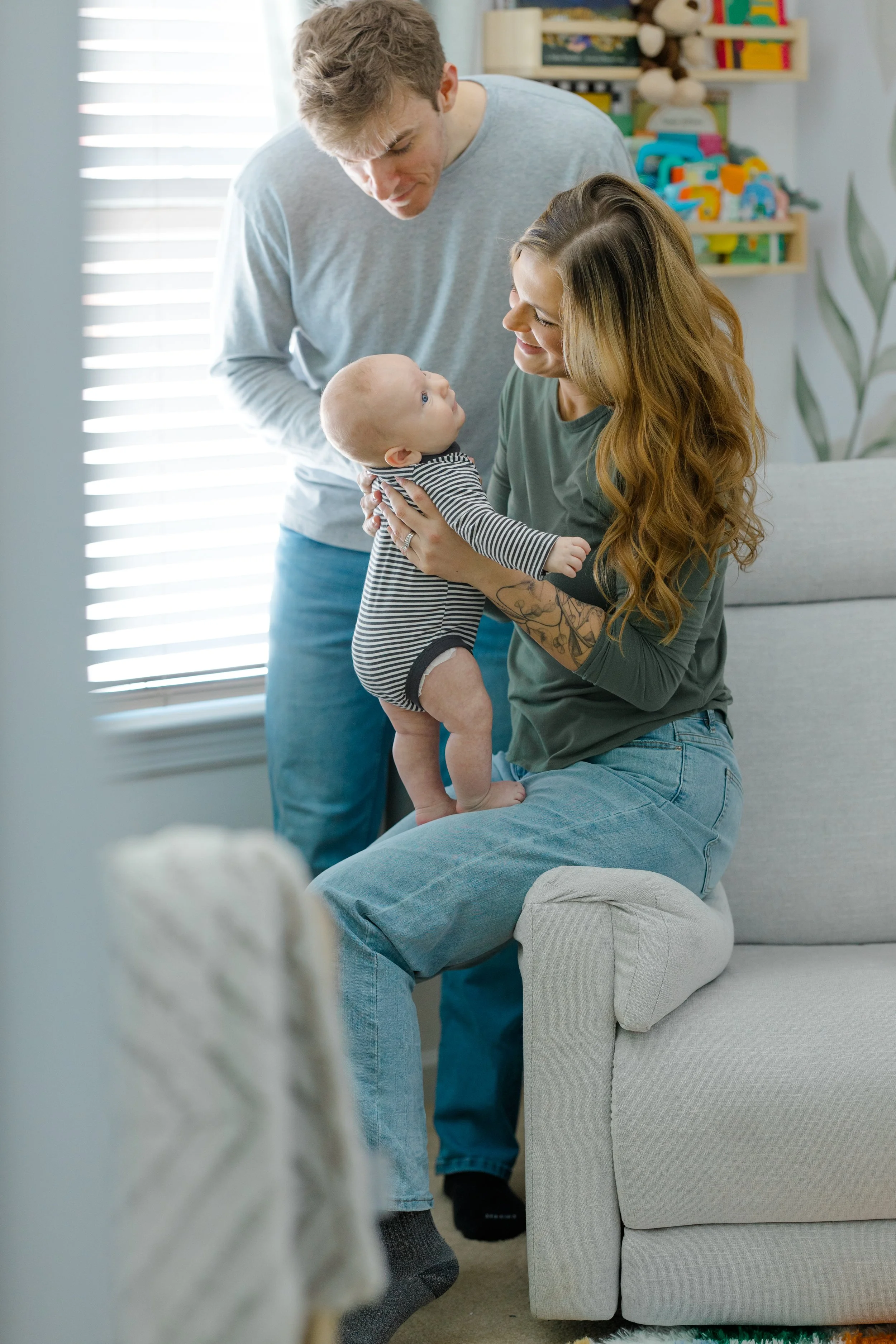 A family of three, with a man and woman and a baby, is sitting together in a well-lit room. The woman, with long wavy red hair, is sitting on a couch holding the baby, who is looking up at her. The man, standing close by, is leaning toward the woman 