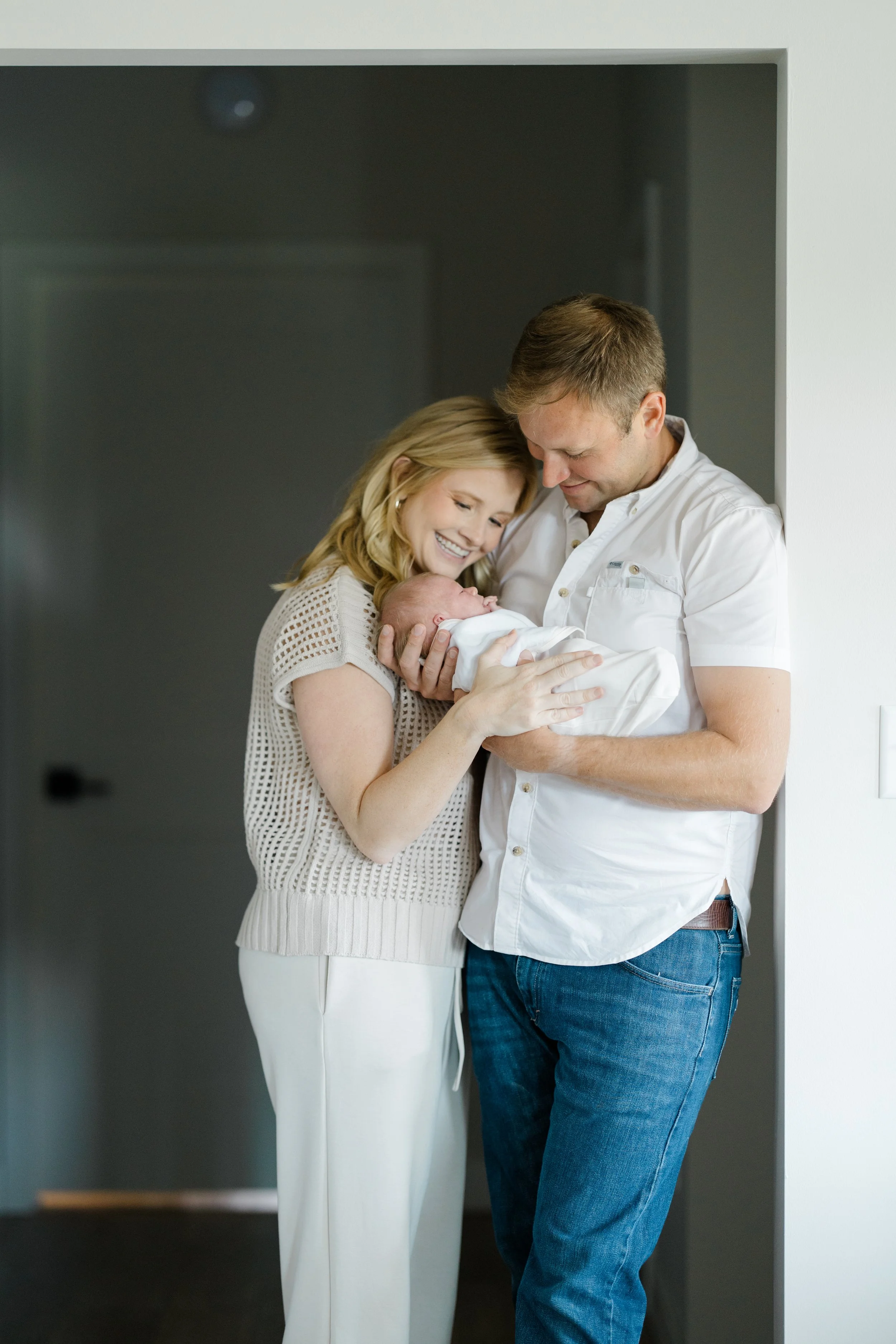 A couple holding a newborn baby in a doorway, both smiling and looking at the baby lovingly.