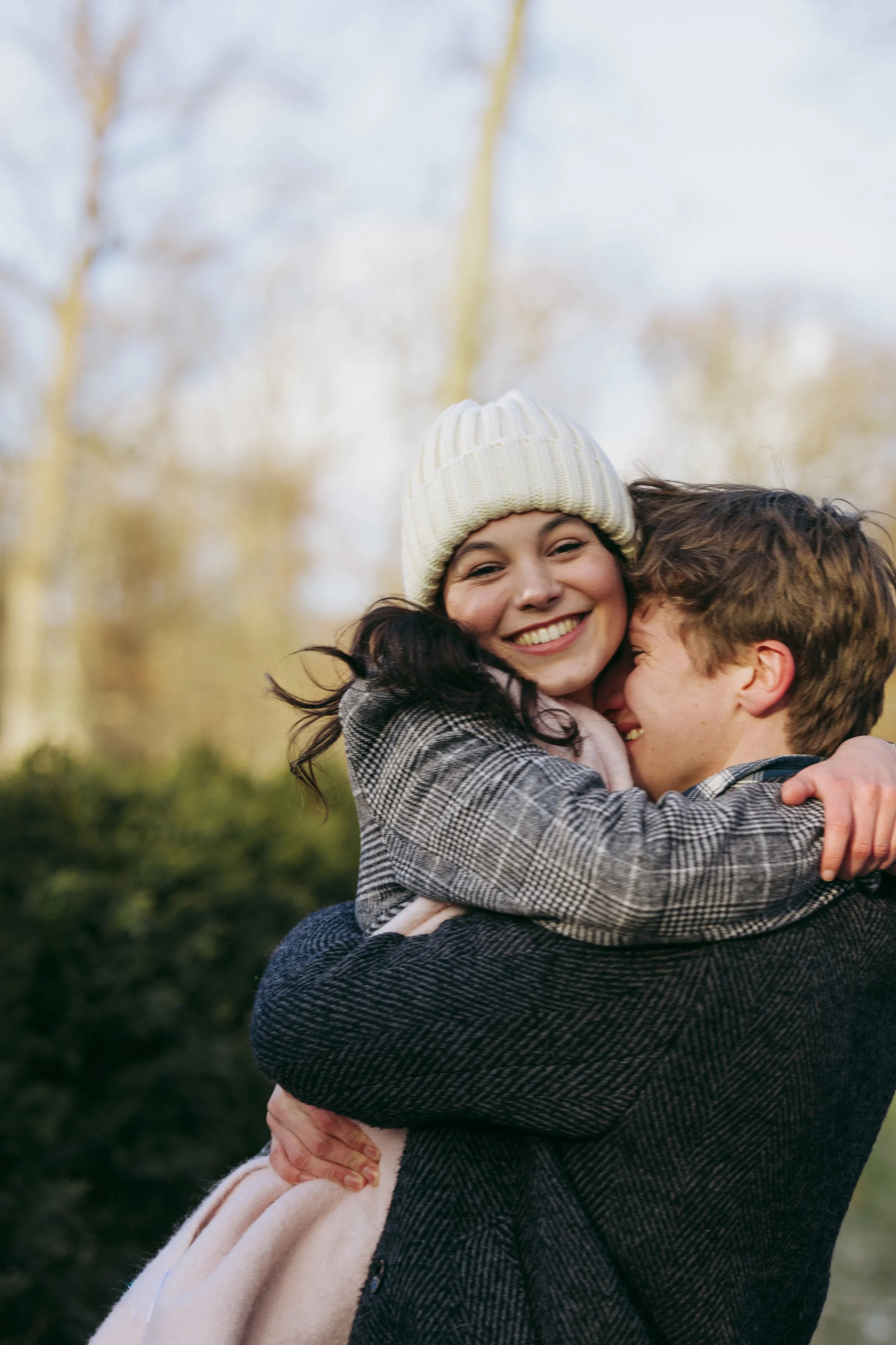 A woman with a white knit hat smiling while hugging a man outdoors on a sunny day with trees in the background.