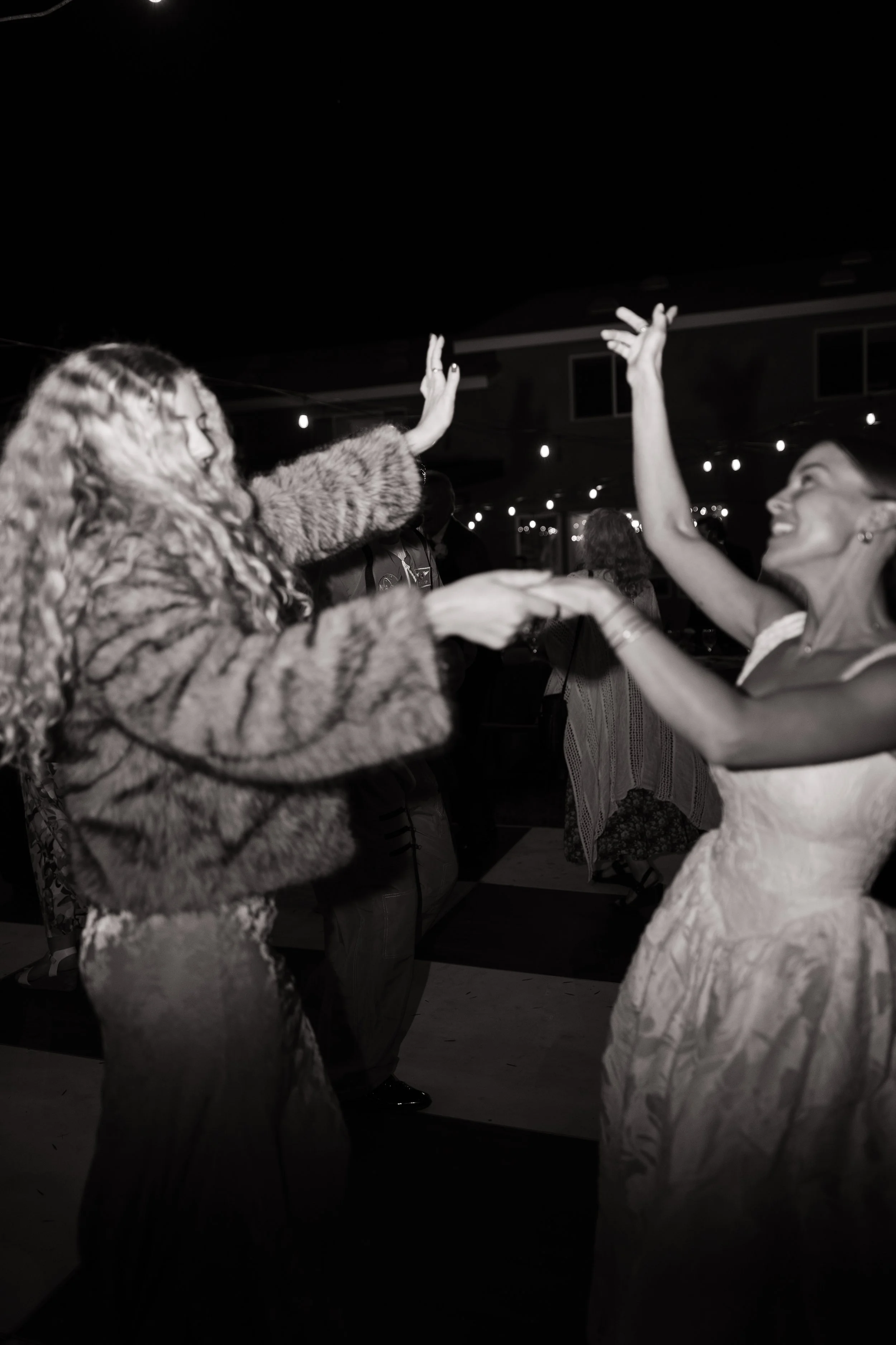 Two women dancing and smiling at a nighttime outdoor event, one wearing a fur coat and the other a dress, with string lights overhead.