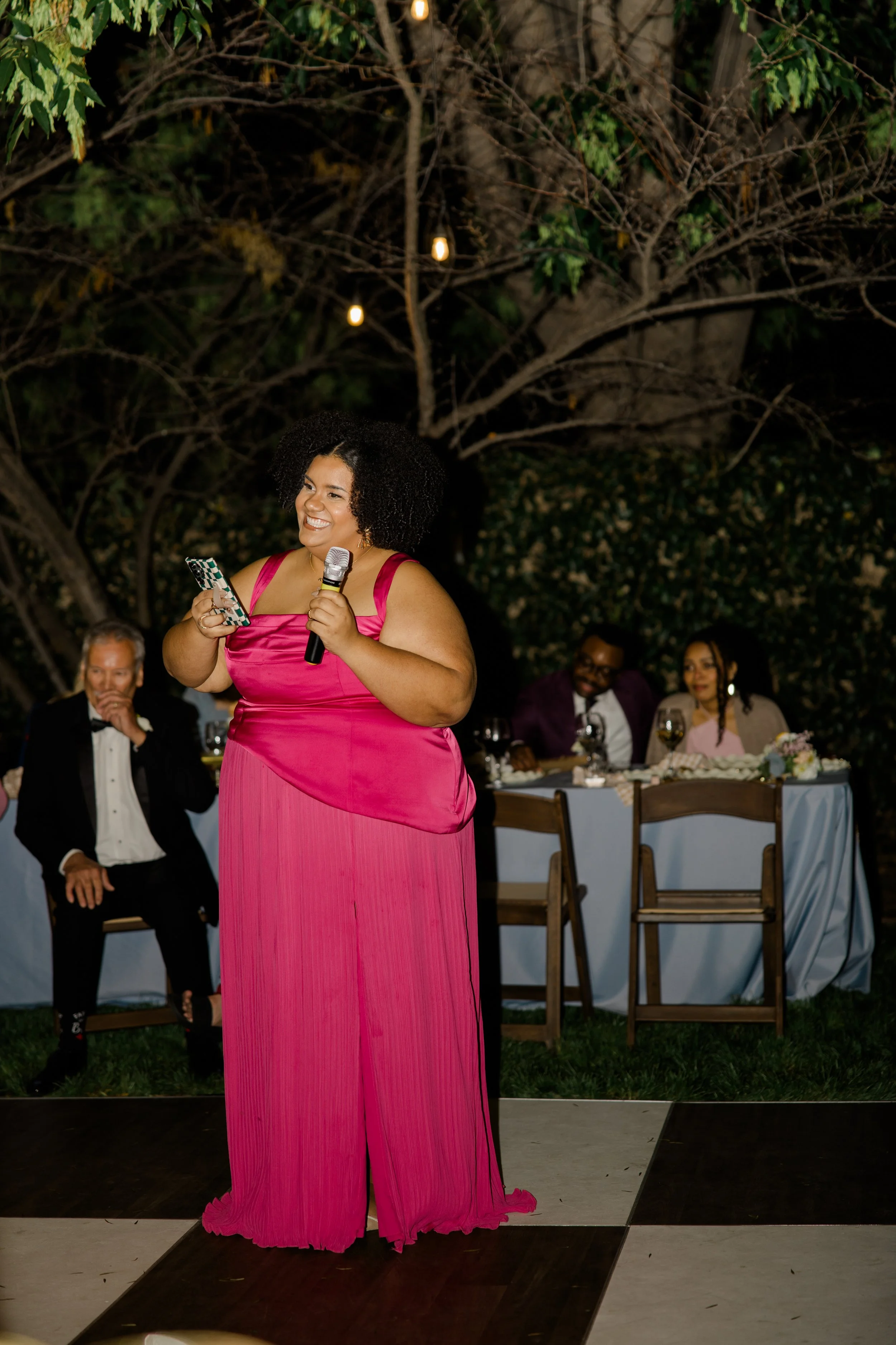 A woman in a pink gown holding a microphone and a phone, standing at an outdoor event during nighttime with trees and people seated at tables in the background.
