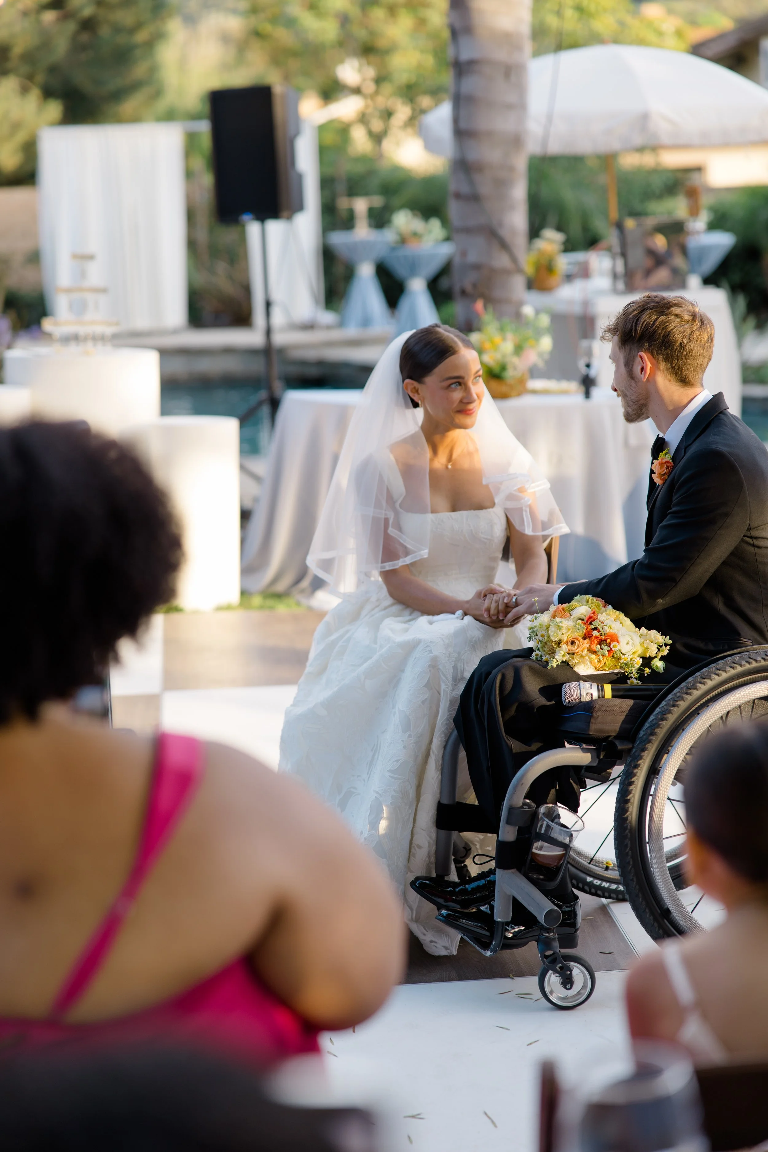 A bride and groom exchanging vows at an outdoor wedding ceremony, with guests seated nearby, in a garden setting with tables, flowers, and an umbrella in the background.