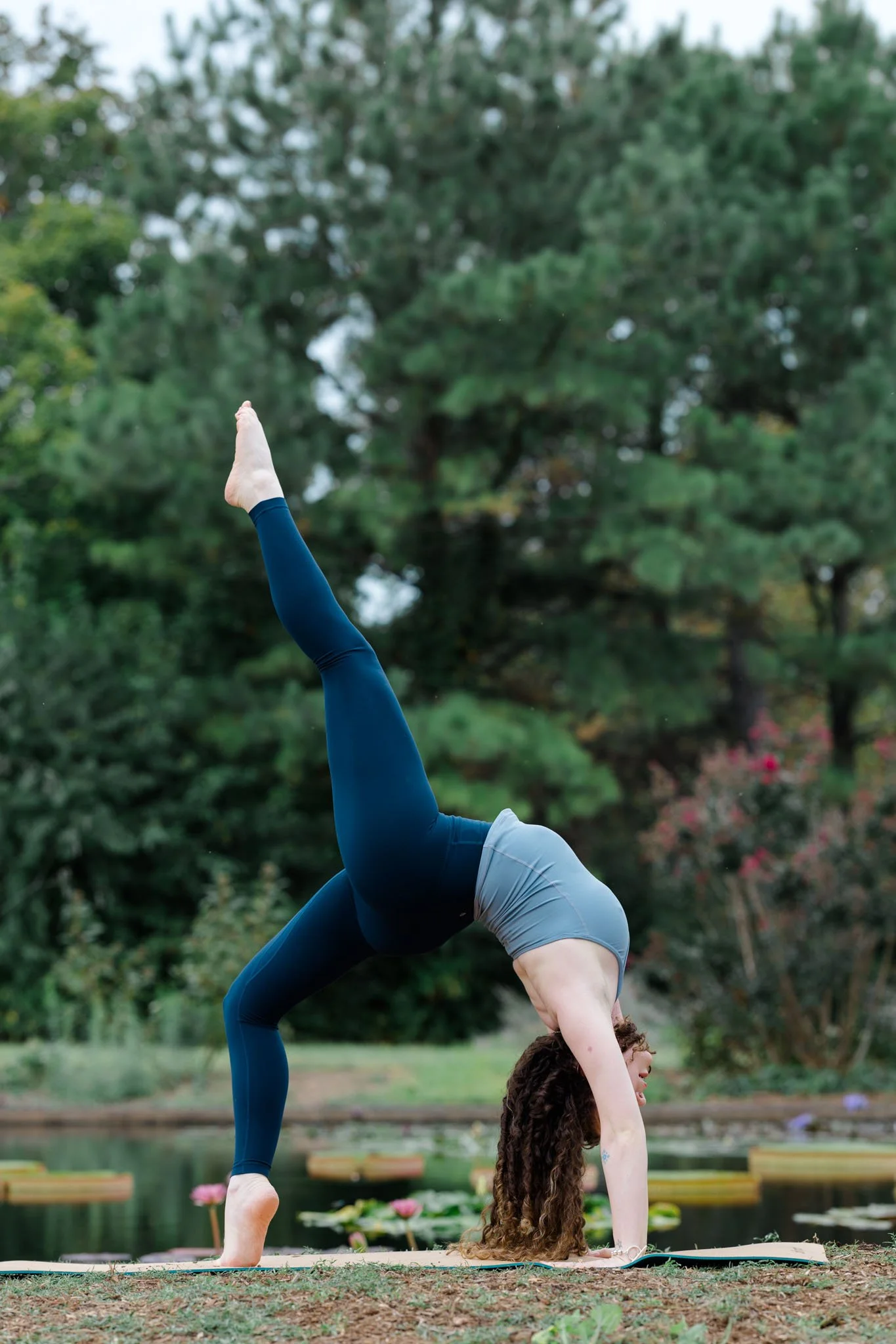A woman practicing yoga outdoors, performing a handstand on a yoga mat in a park with trees and water lilies in the background.