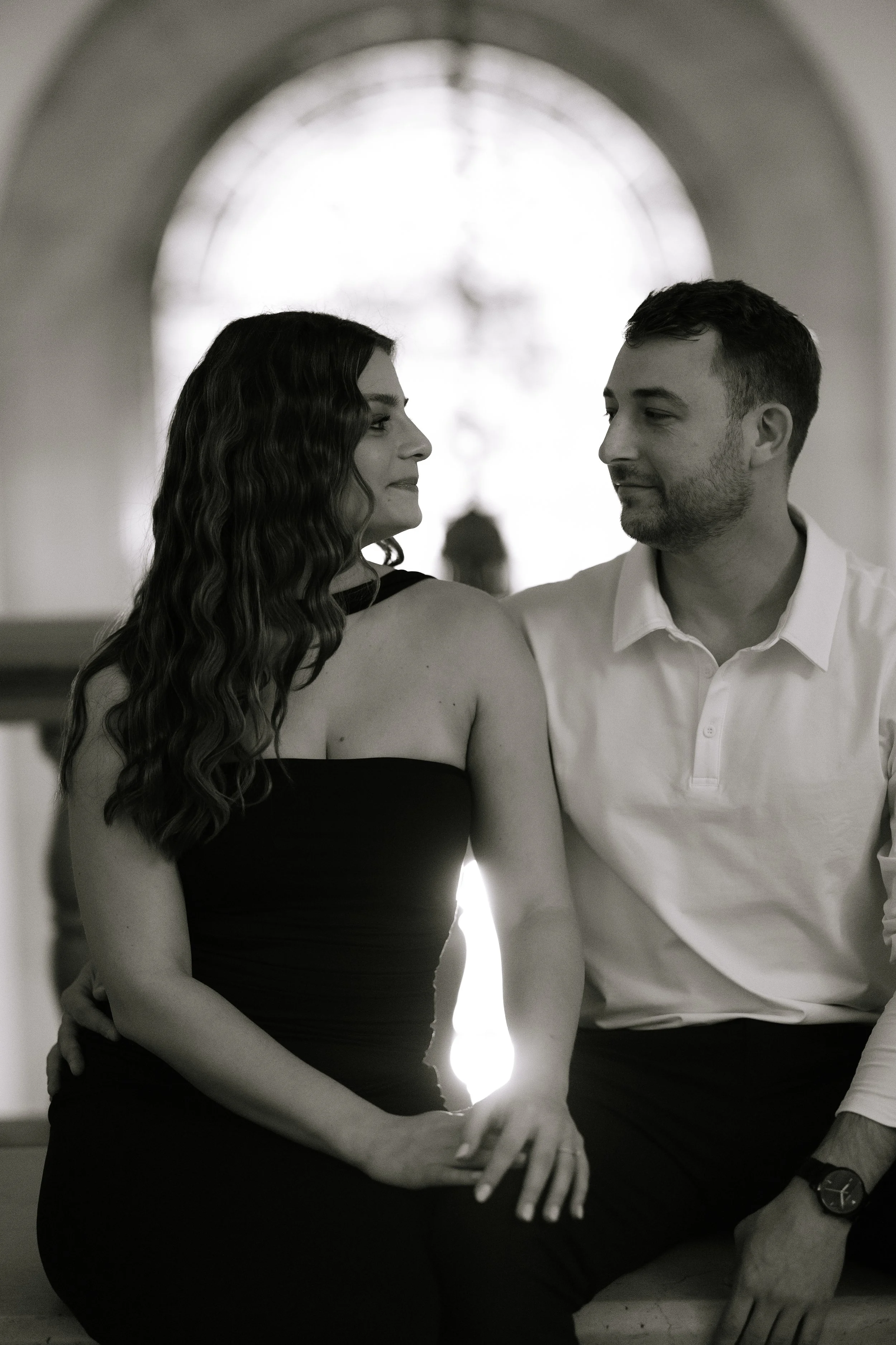 A black and white photo of a man and a woman sitting close together indoors, with a large window or light source behind them. The woman has long wavy hair and is wearing a strapless black top, while the man has short hair, a slight beard, and is dres