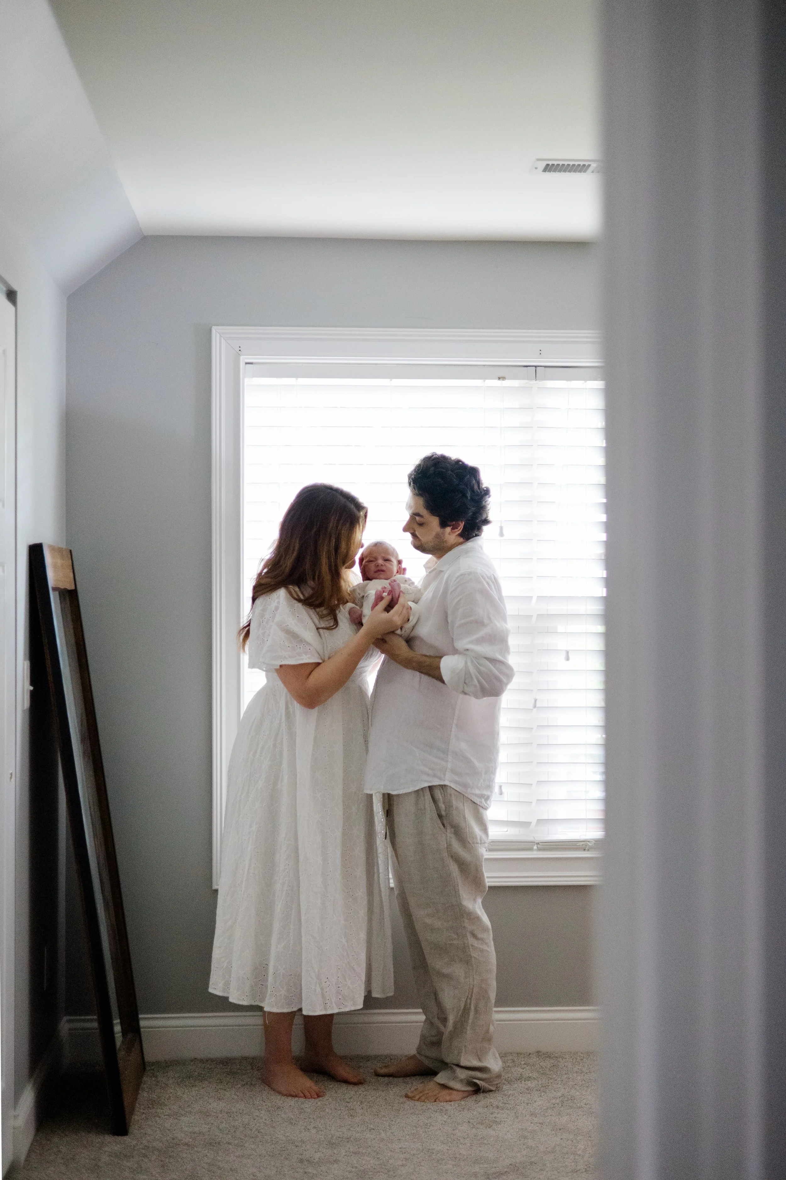 A couple holding a baby, standing barefoot inside a room near a window with white blinds, with gray and white walls and a carpeted floor.