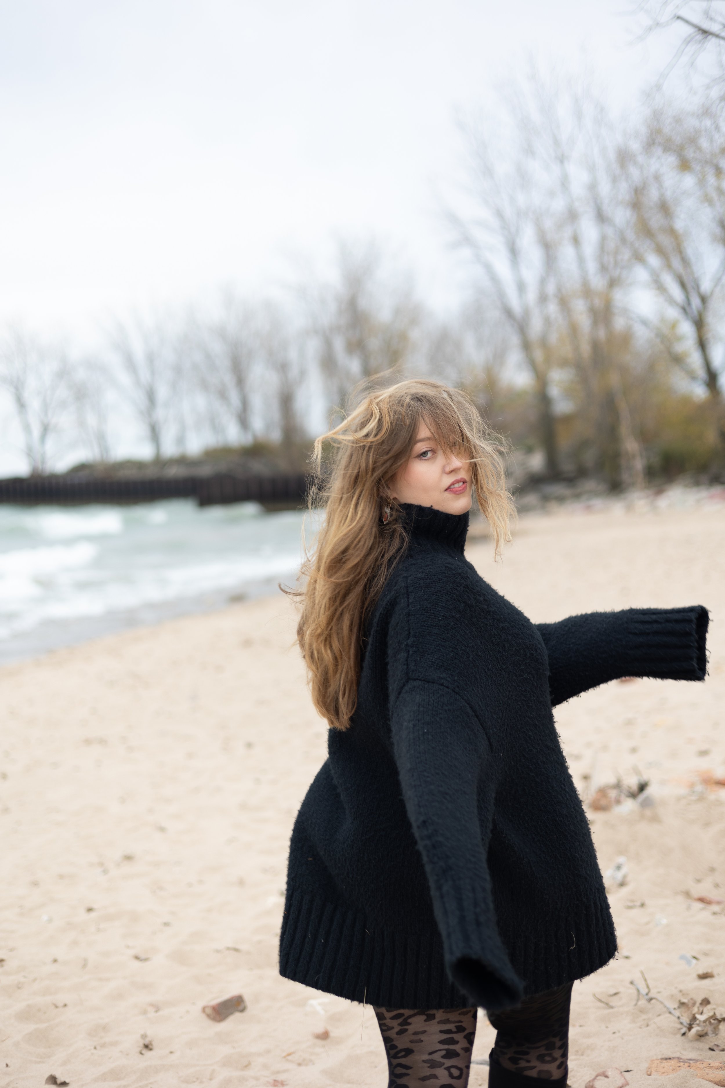 A young woman with long wavy hair and a nose piercing wearing a black sweater, standing on a sandy beach with trees in the background during overcast weather.