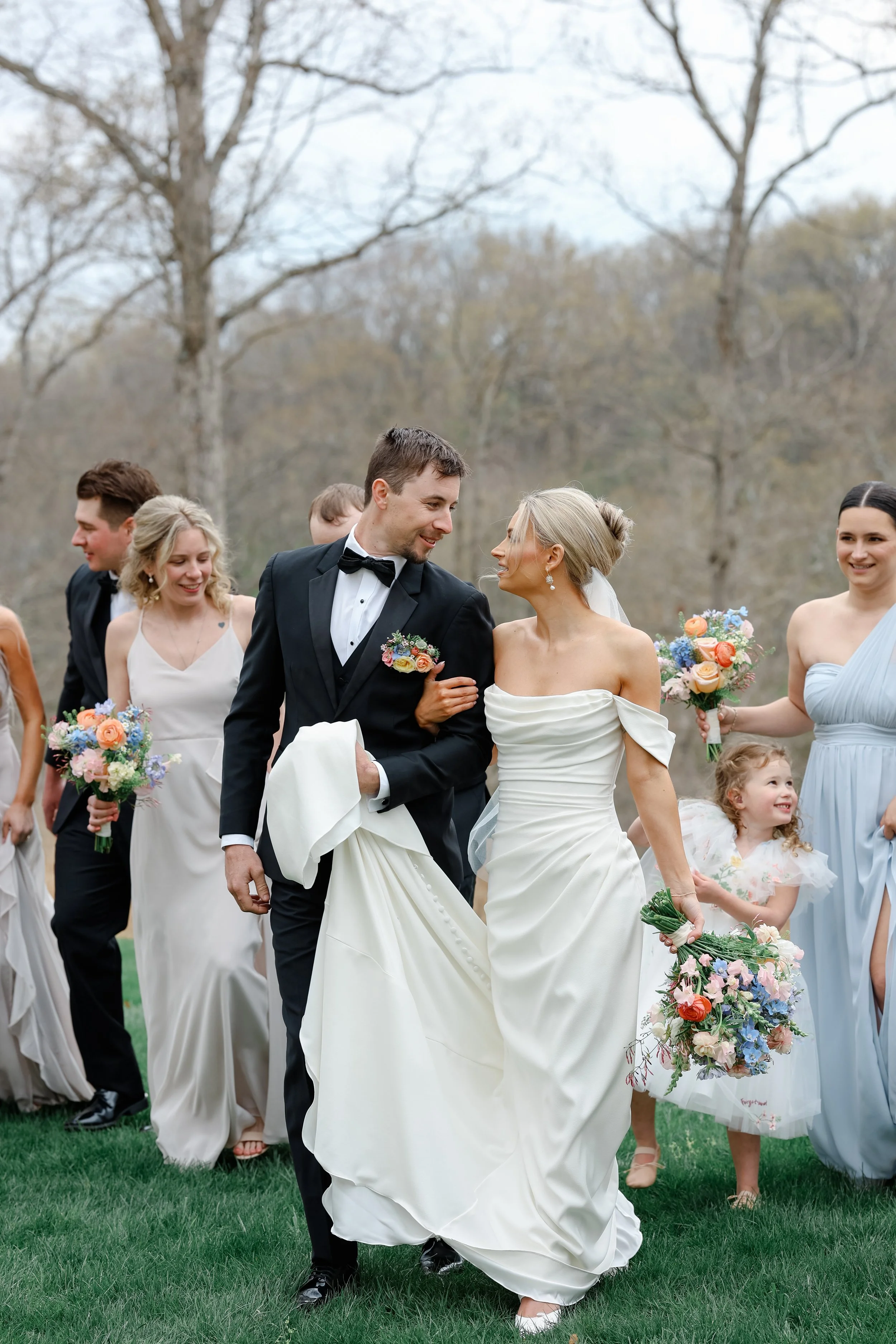 A bride and groom walking arm-in-arm outdoors during a wedding celebration, surrounded by bridesmaids and bridesmen holding bouquets, with trees and a cloudy sky in the background.