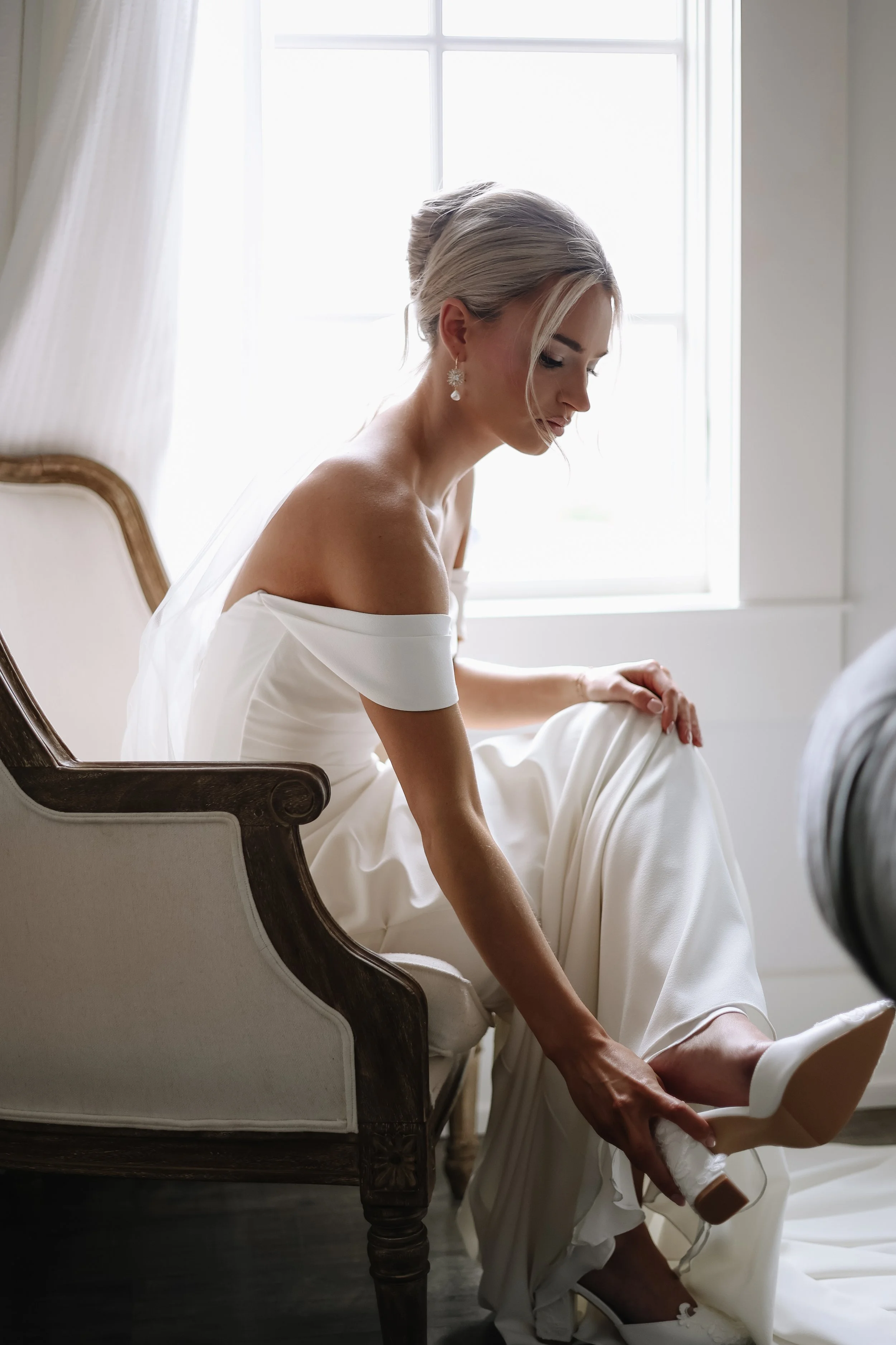 A woman in a white dress sitting on a vintage chair near a window, adjusting her shoe as she leans forward.