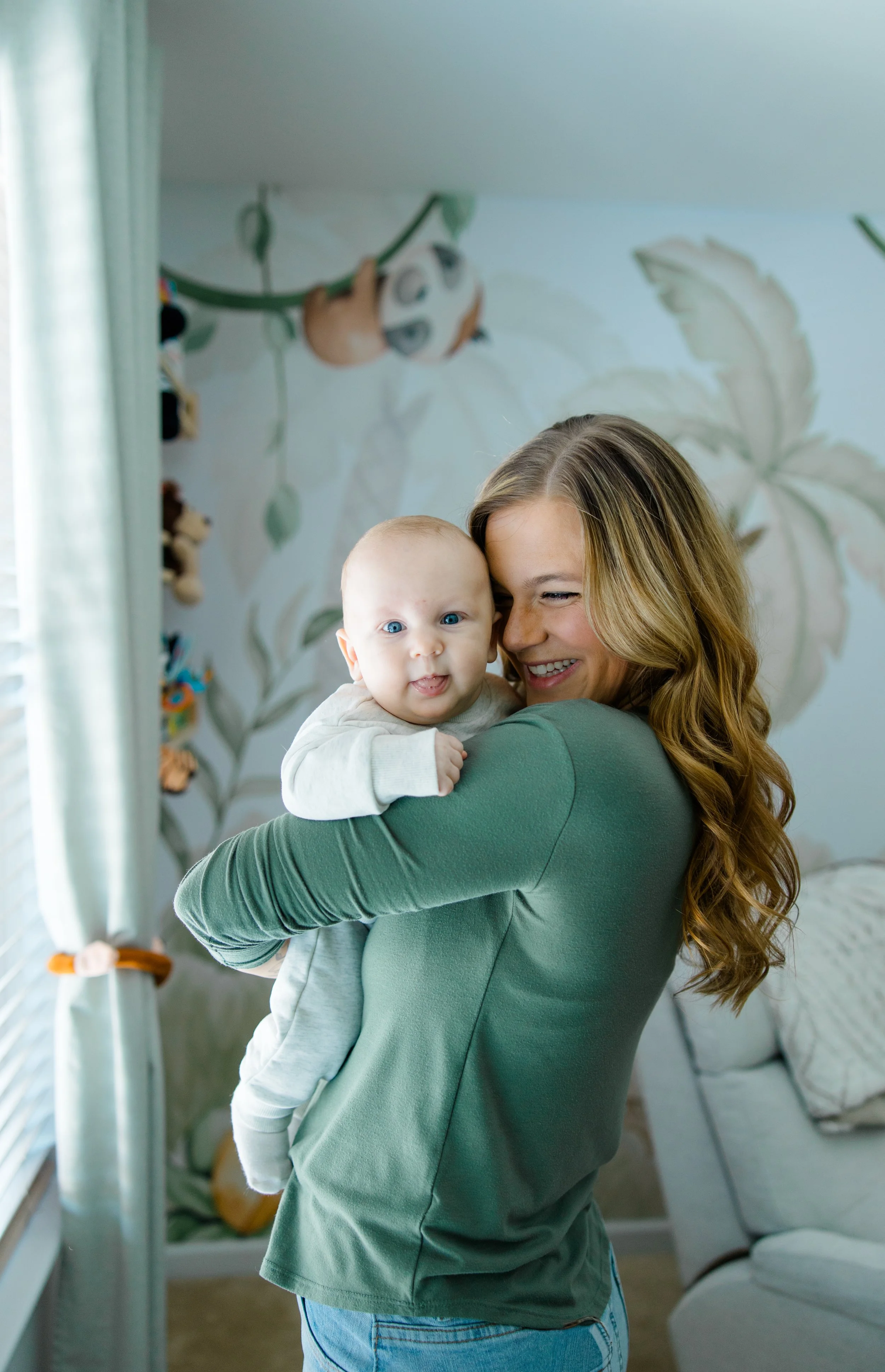 A woman with long wavy blonde hair smiling and holding a baby with light hair and blue eyes indoors near a window with curtains. There are stuffed animals hanging from the ceiling and a bed with a sconce on the wall in the background.