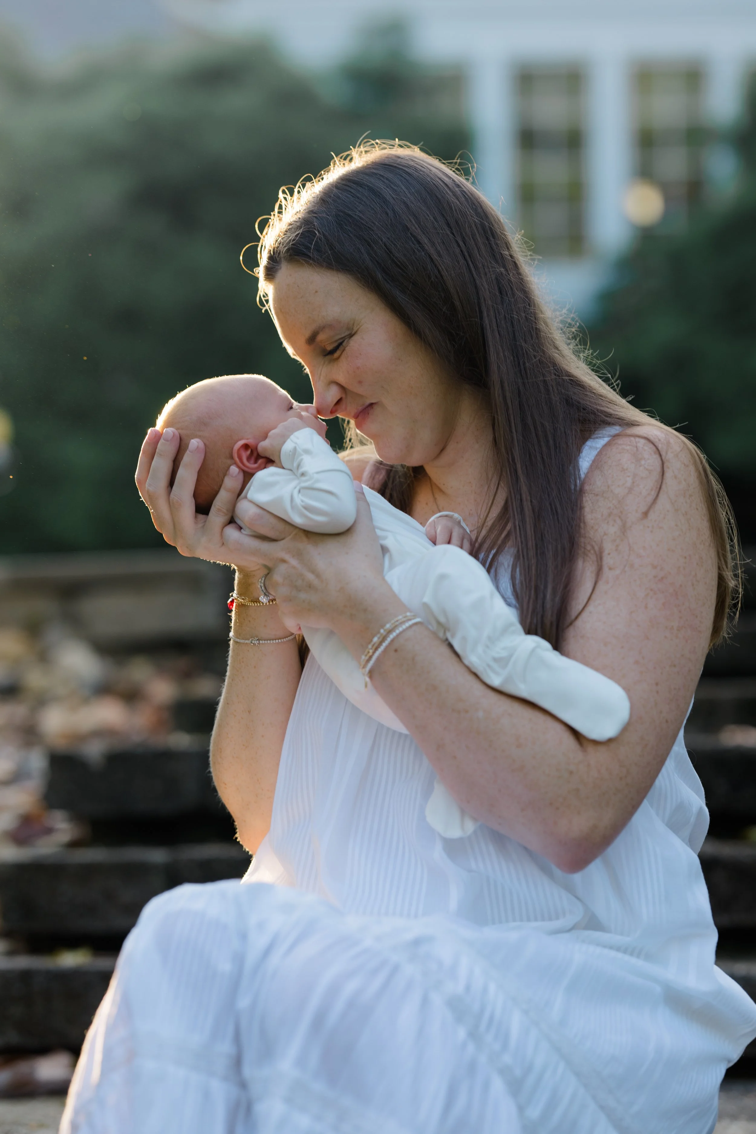 A woman with long brown hair and wearing a white dress is holding a baby with a bald head and light skin. The woman is smiling and touching her forehead to the baby's. The background is outdoors, blurred, with lush greenery and steps.