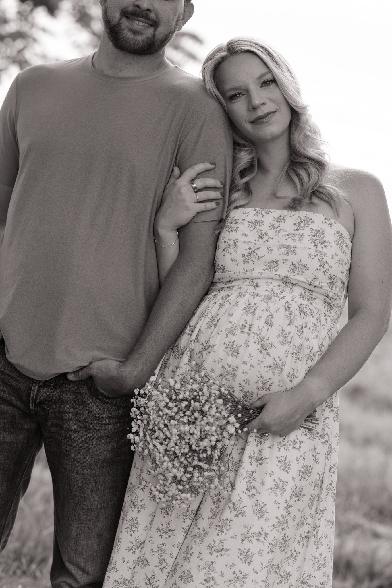 A black and white photo of a couple standing close together outdoors, with the woman resting her head on the man's shoulder, holding a bouquet of small flowers.