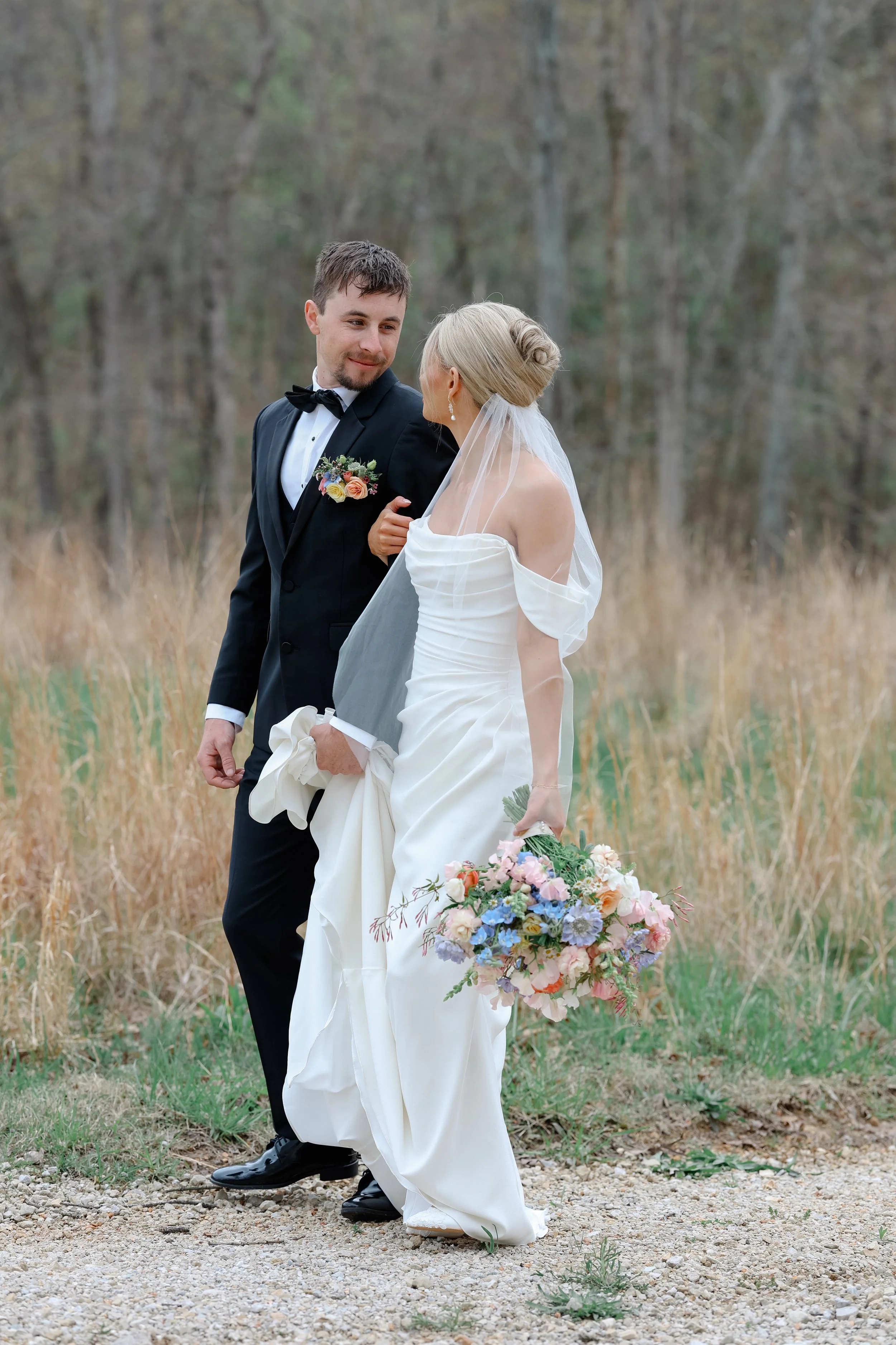 A bride and groom walking outdoors in a field with trees in the background. The bride is holding a colorful bouquet of flowers and the groom is wearing a black tuxedo with a boutonniere.