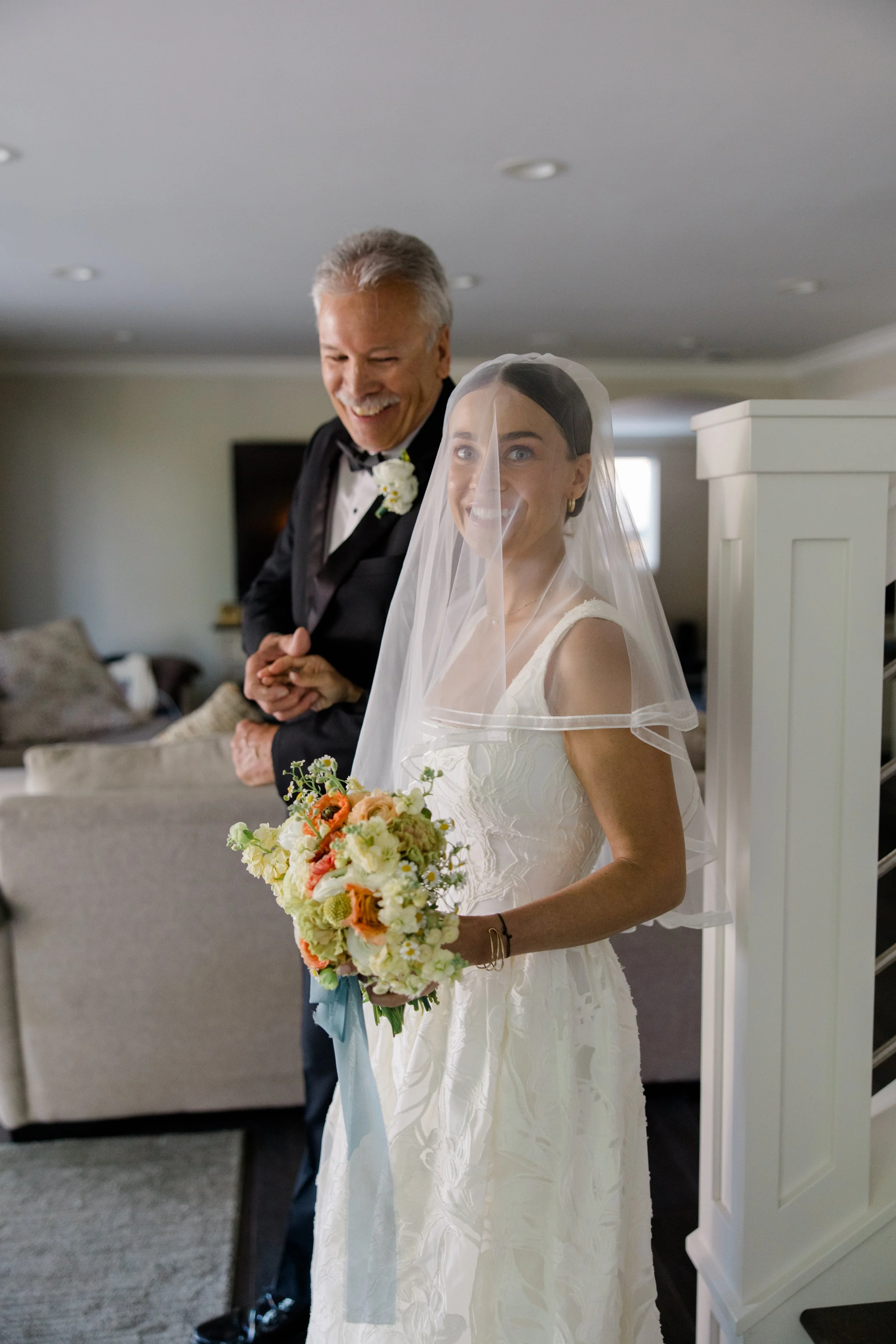 A bride is smiling and holding a bouquet, with a man in a tuxedo in the background, inside a room with neutral decor.