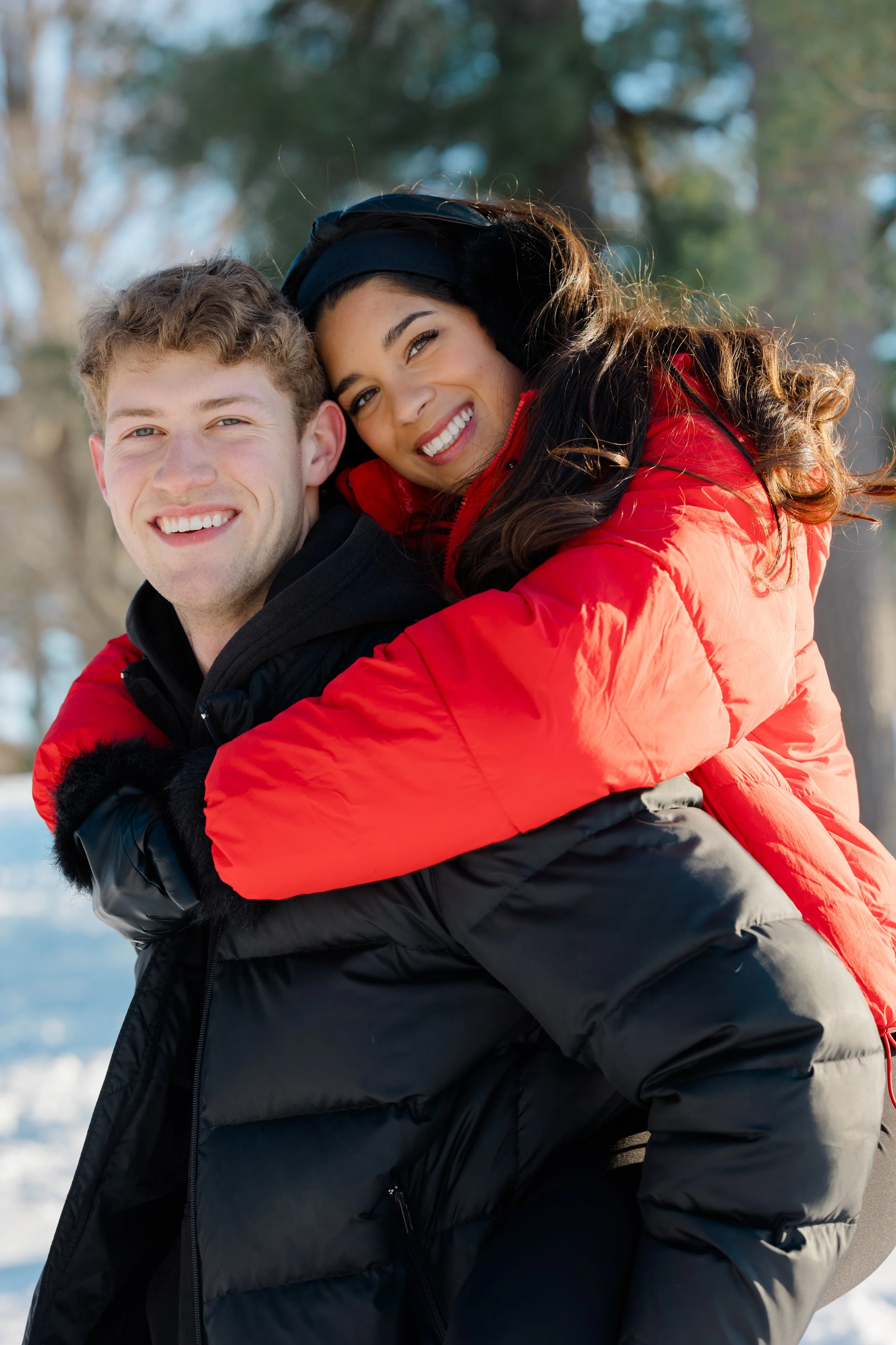A smiling young man with curly hair and a young woman with long brown hair, both wearing winter jackets, engage in a joyful piggyback ride outdoors in a snow-covered area with trees in the background.