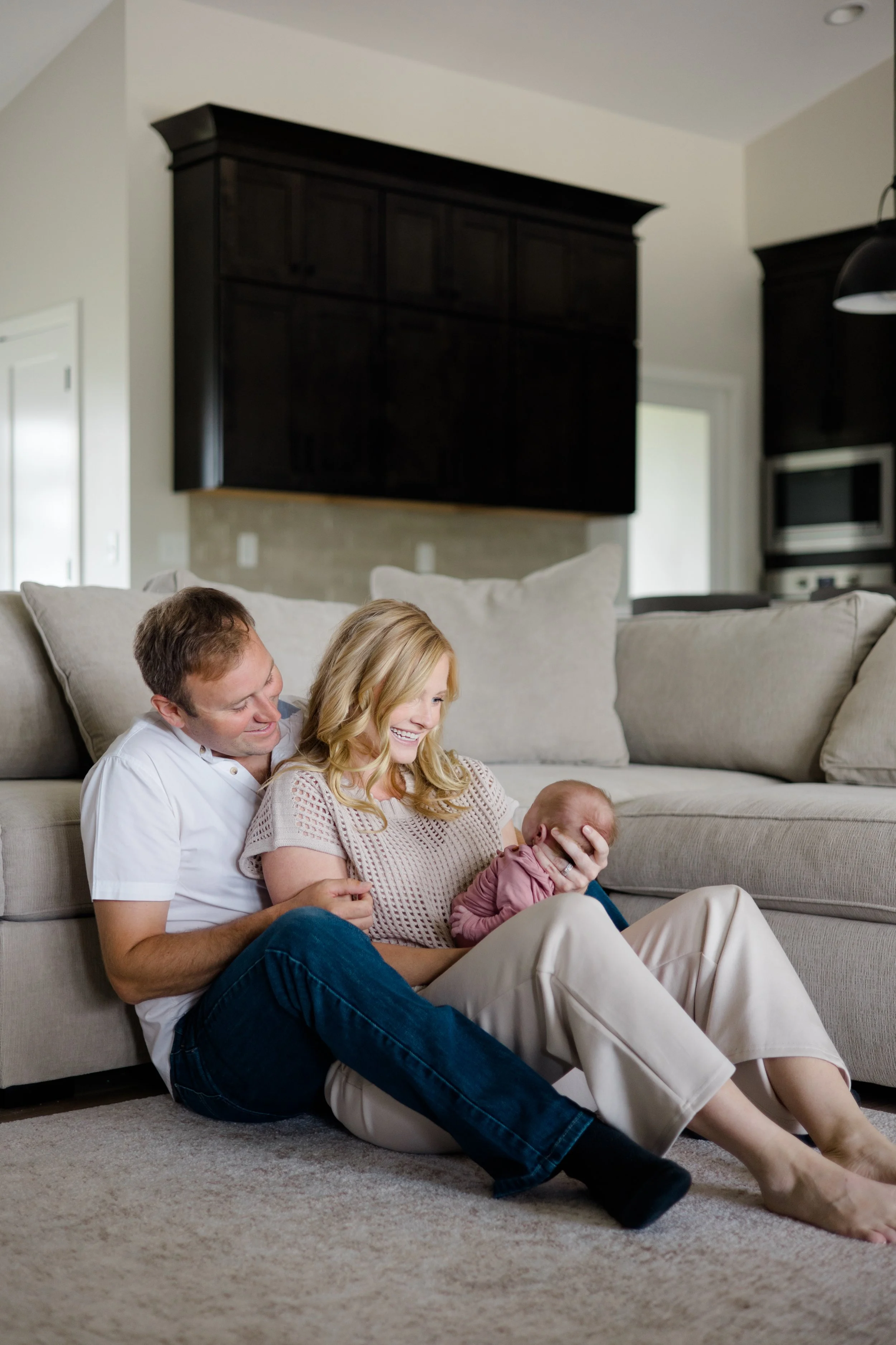 A family of four sitting on the floor and a beige couch in a living room. The father, mother, and a young child are sitting on the floor, smiling and looking at a newborn baby the mother is holding.
