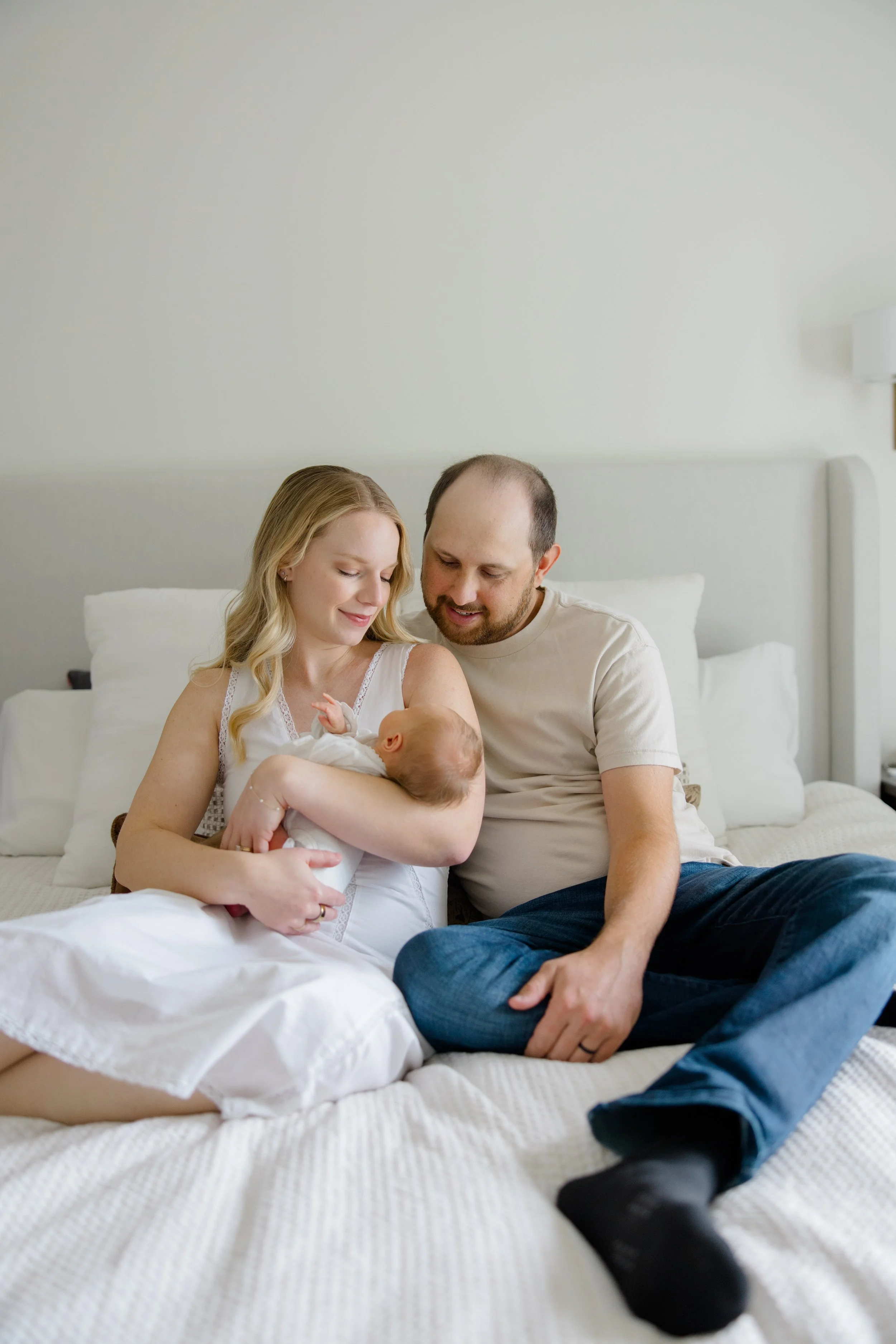 A woman and a man sitting on a bed with a newborn baby. The woman is holding the baby and smiling, while the man is looking at the baby with a smile. The setting is a bedroom with a light-colored wall and pillows in the background.