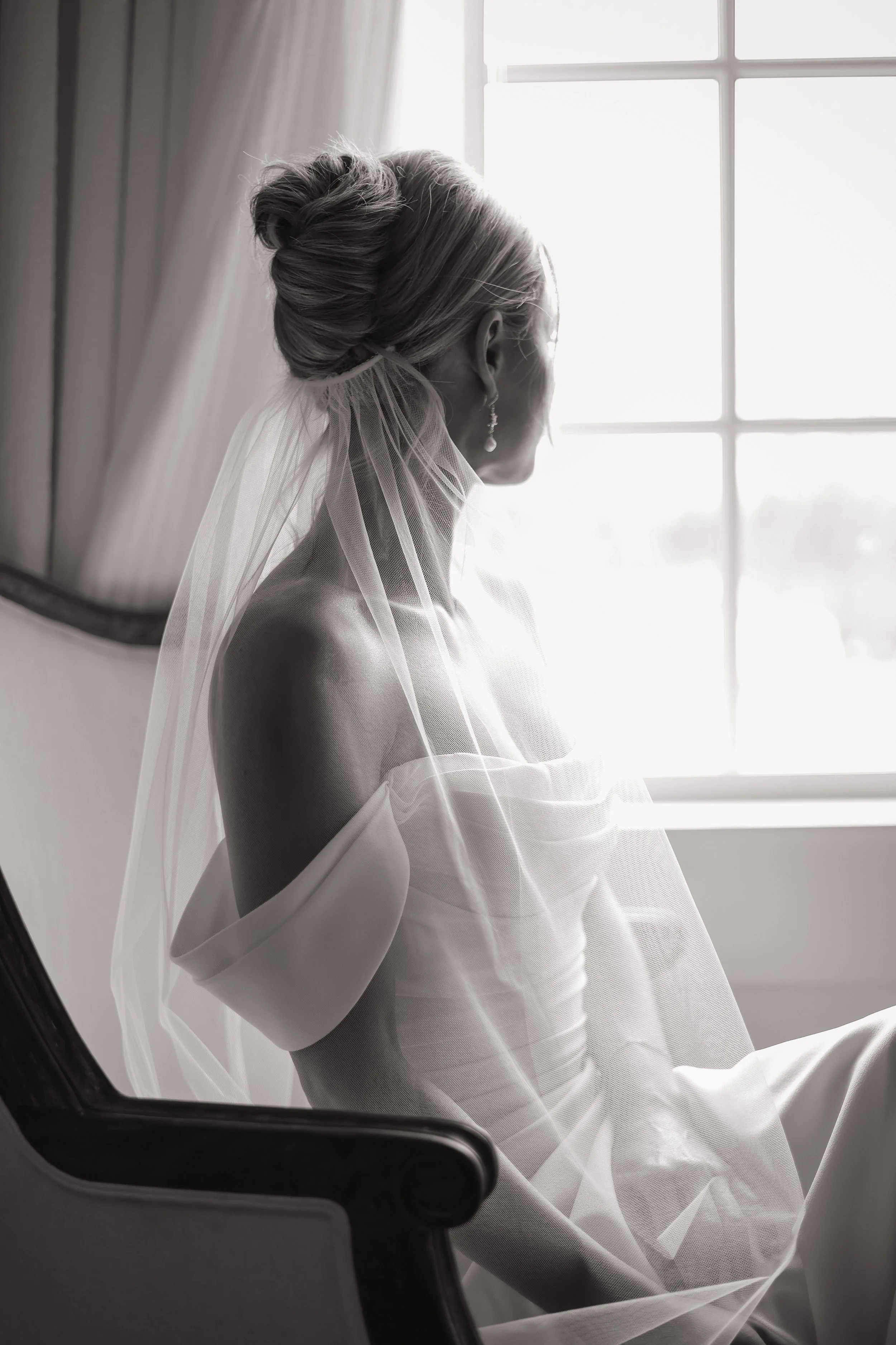 Black and white photograph of a woman with her hair pinned up, gazing out a window with sunlight streaming in, wearing a veil and earrings.